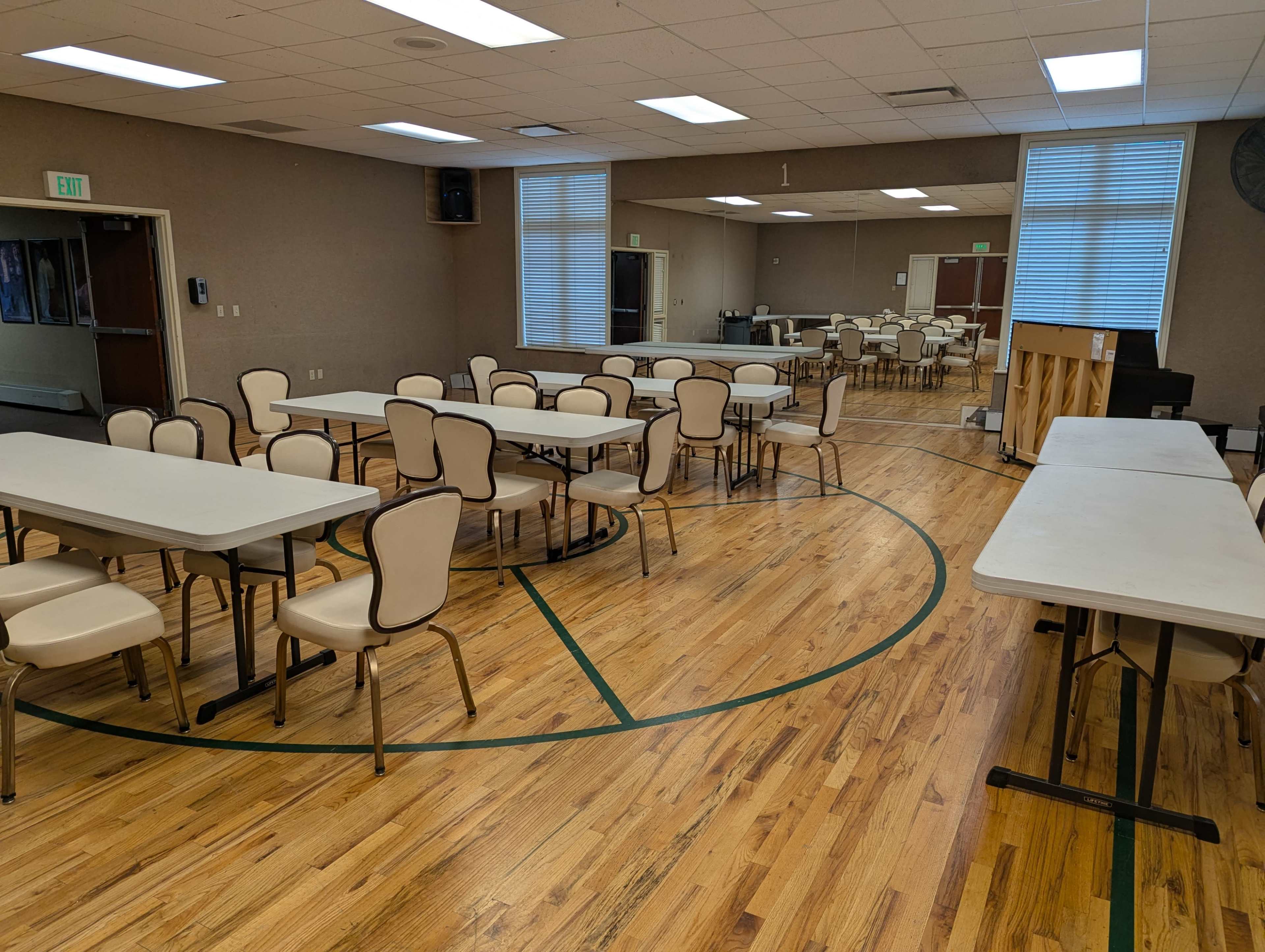 The image shows a spacious community hall with multiple rows of round and rectangular tables surrounded by beige chairs on a wooden floor.