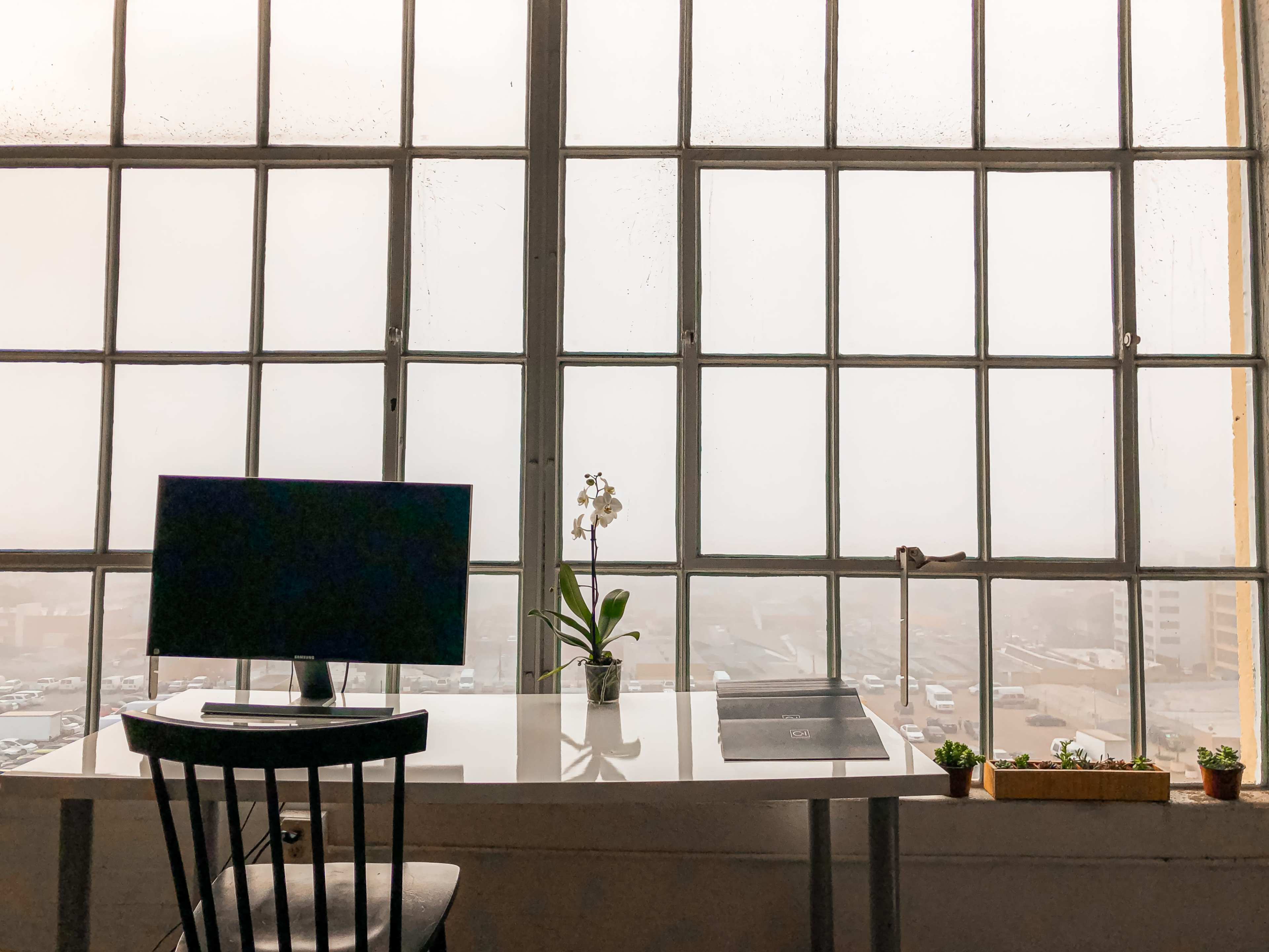 A desk with a computer is positioned near a large window with multiple panes, overlooking a rainy cityscape.