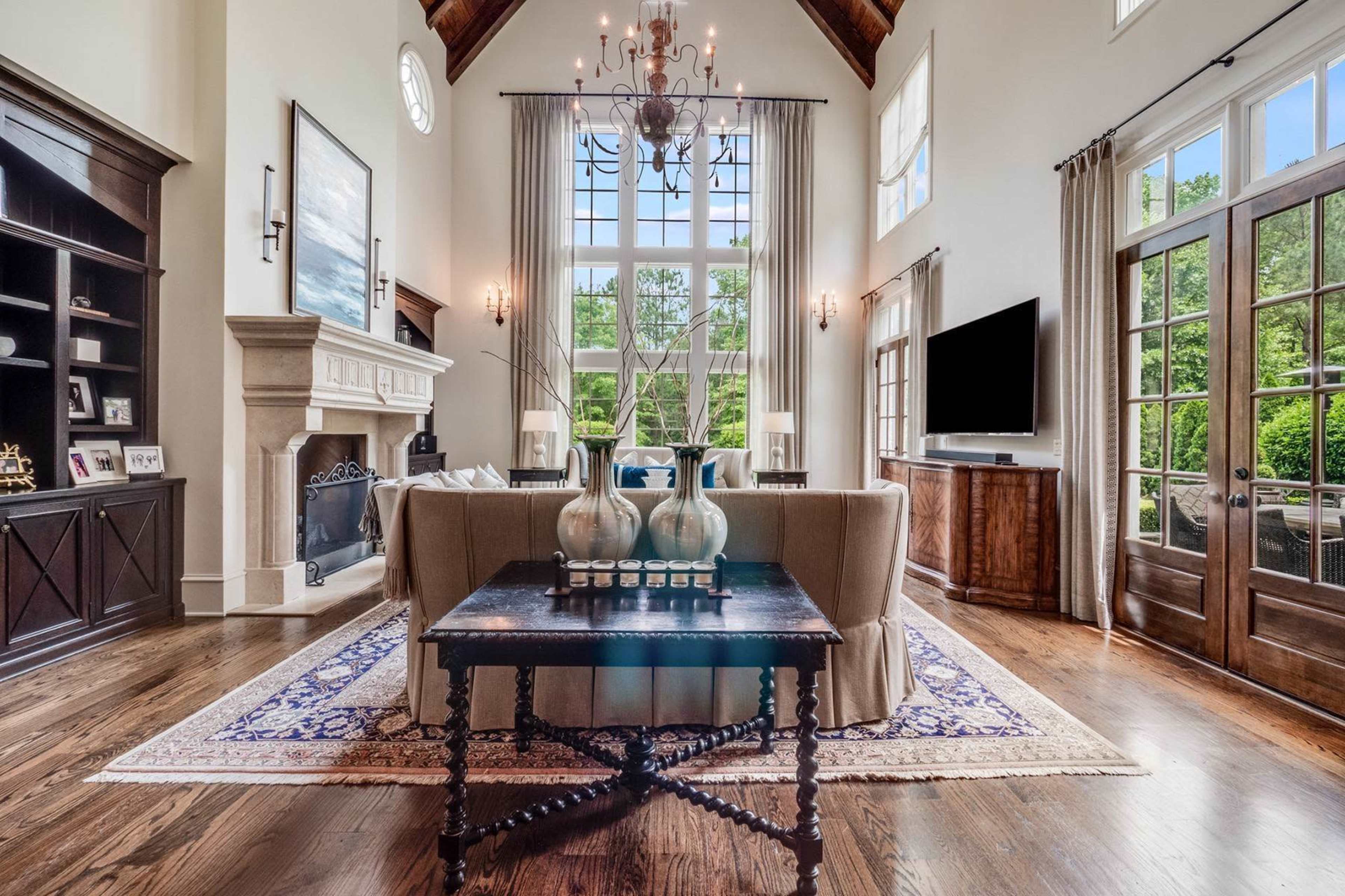 The interior of a spacious living room features a high ceiling, large windows, and elegant furnishings with a coffee table displaying decorative vases.