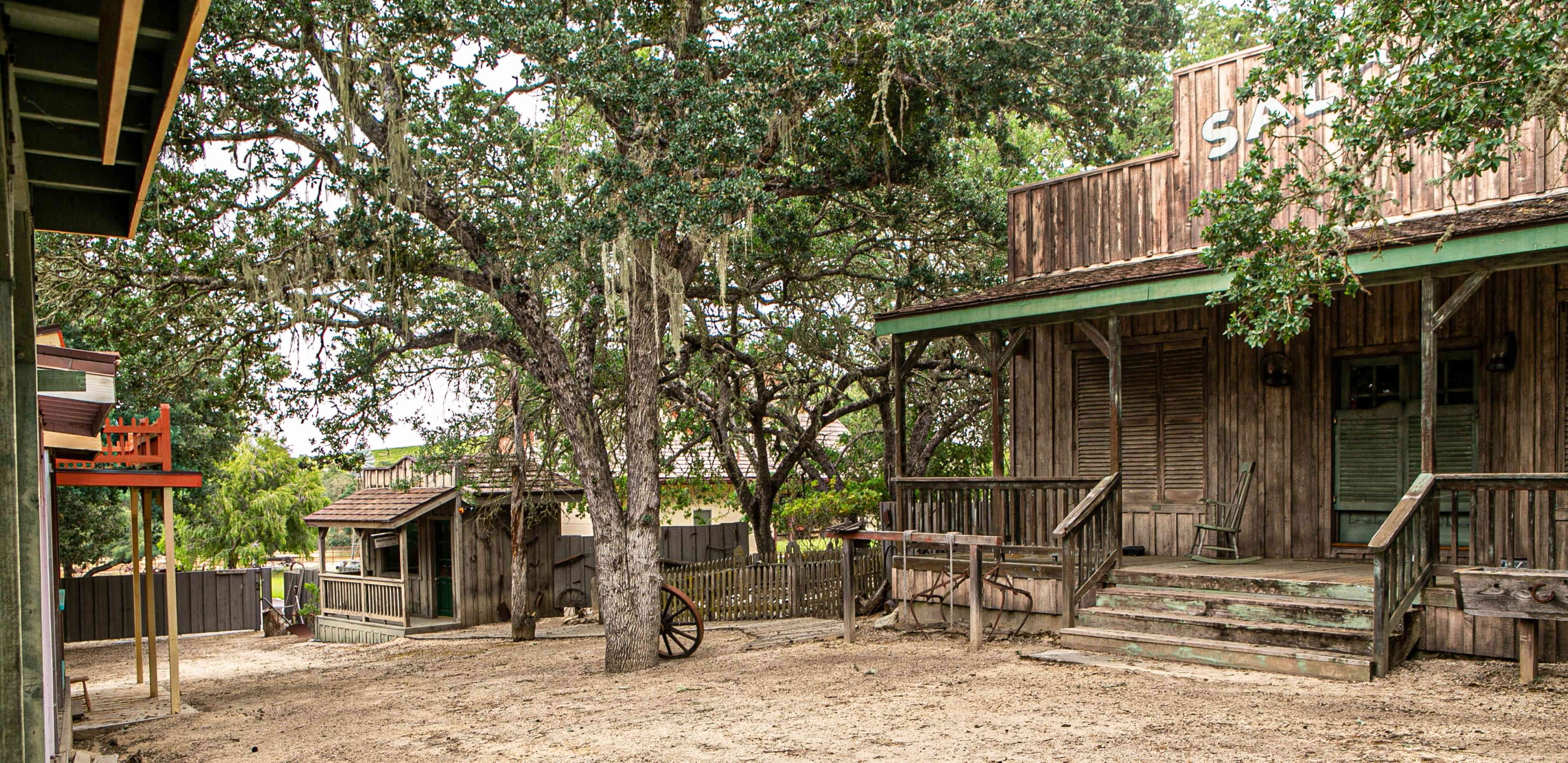 The scene depicts a rustic western-style street with wooden buildings, a sandy area, and large oak trees.