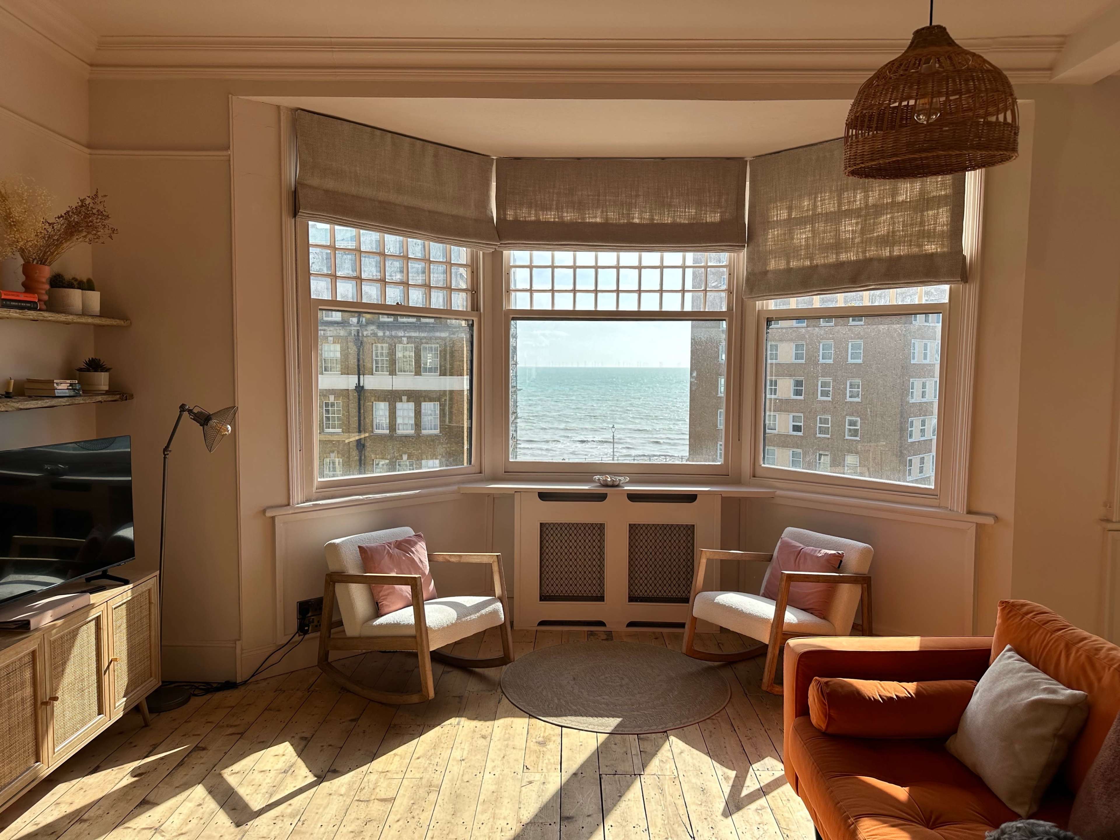 A sunlit living room features two chairs and a sofa, with large windows offering a view of the sea.