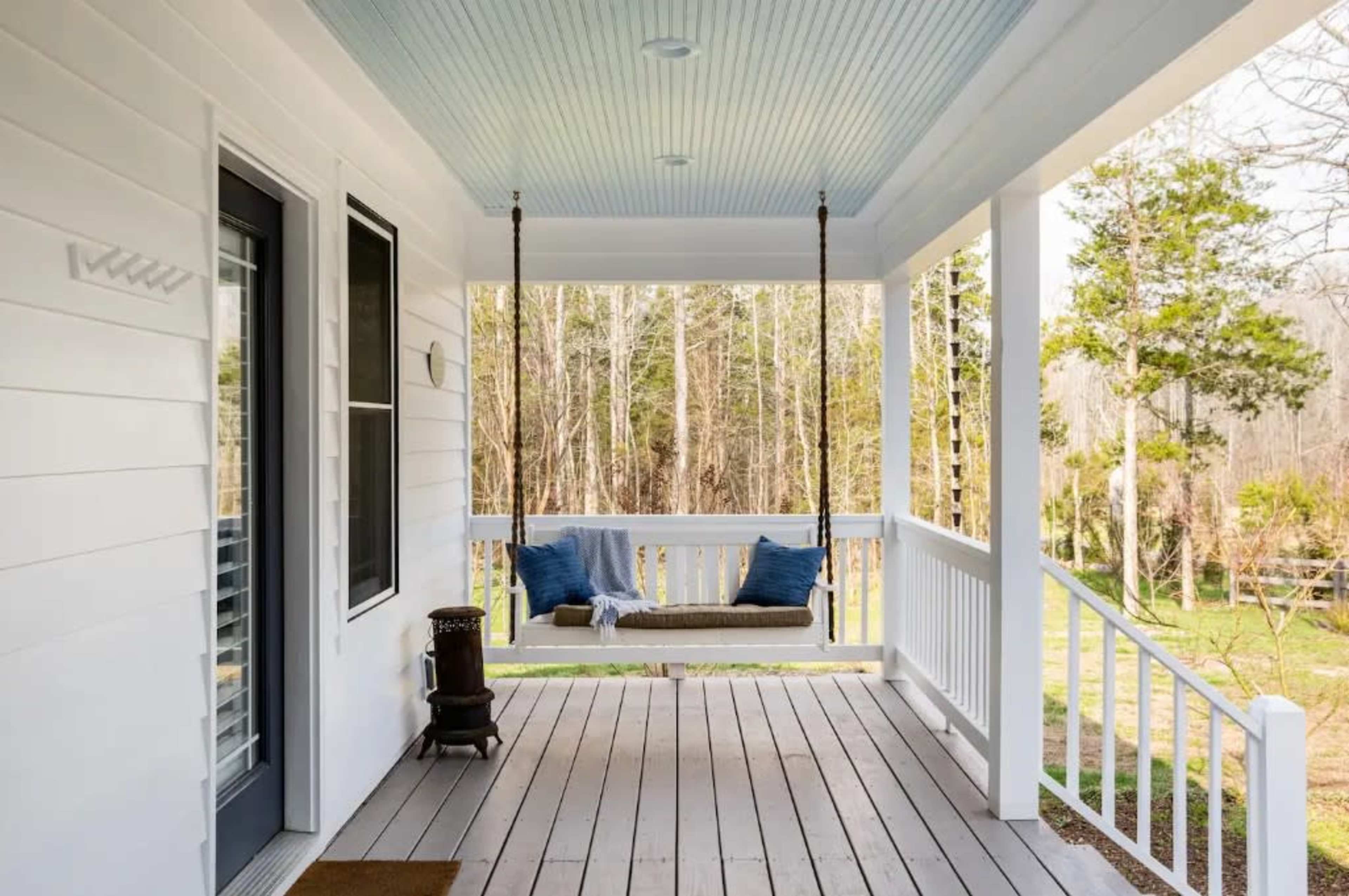 A porch features a swing hanging from the ceiling, two blue pillows on the swing, and a view of a wooded area beyond.