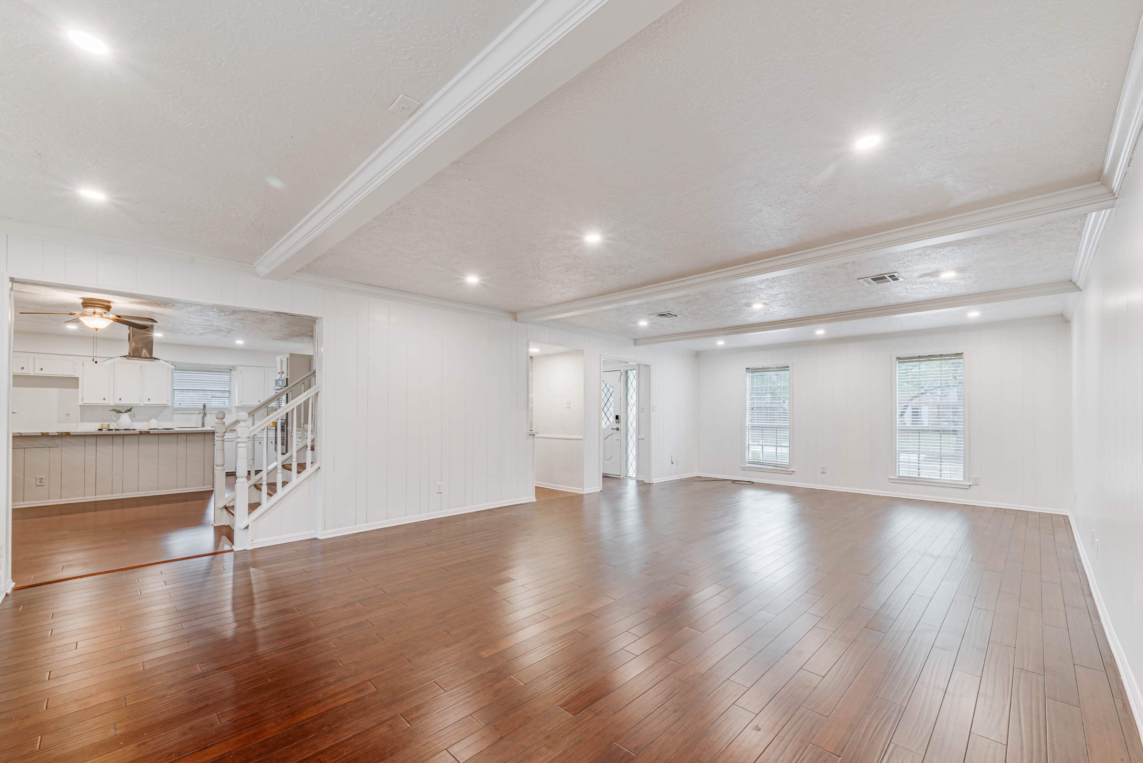 The image shows a spacious, well-lit living room with hardwood flooring and large windows, connected to a staircase and an adjacent kitchen area.