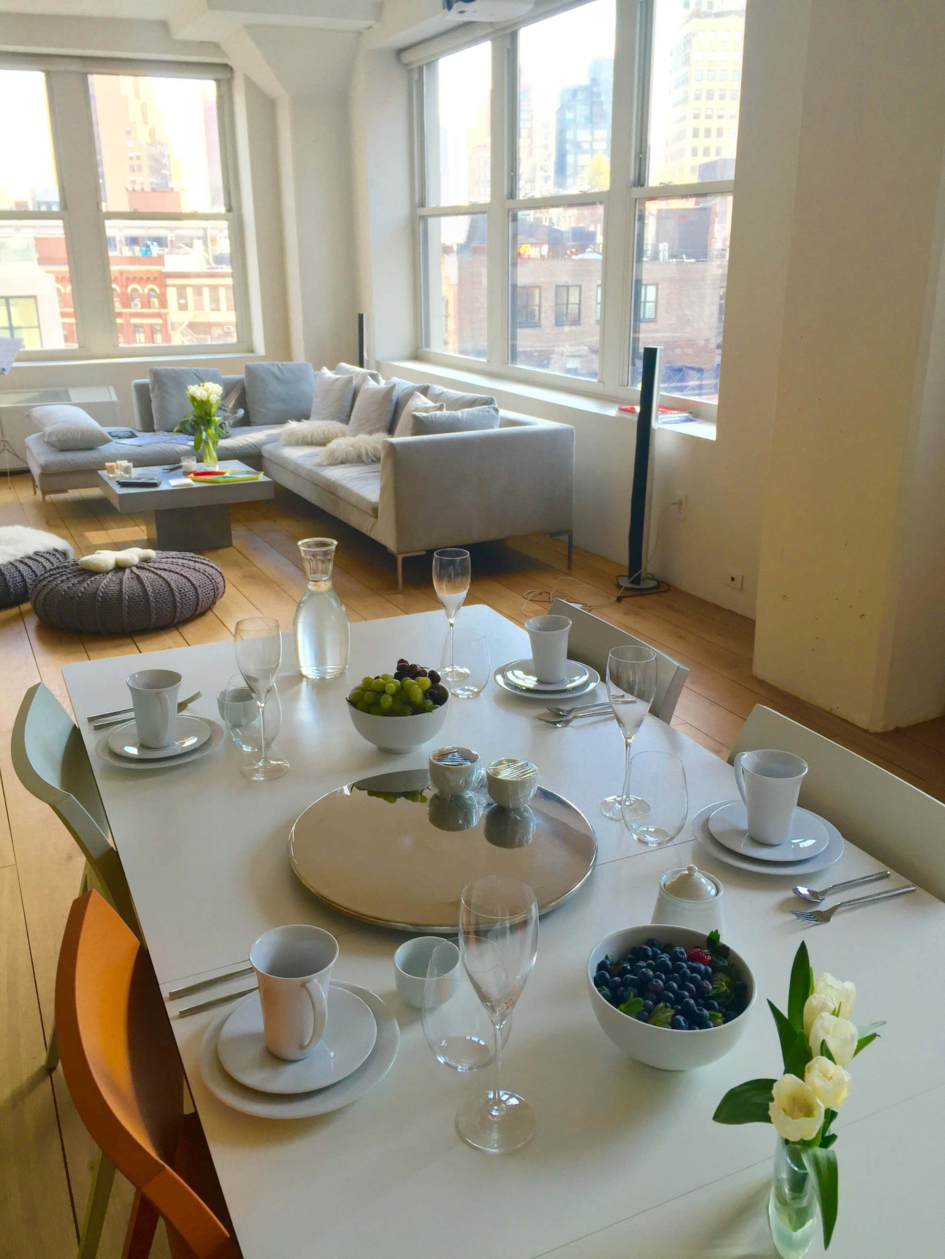 A dining table is set with dishes, glasses, and bowls of fruit, while a seating area with a couch and decorative pillows is visible in the background.