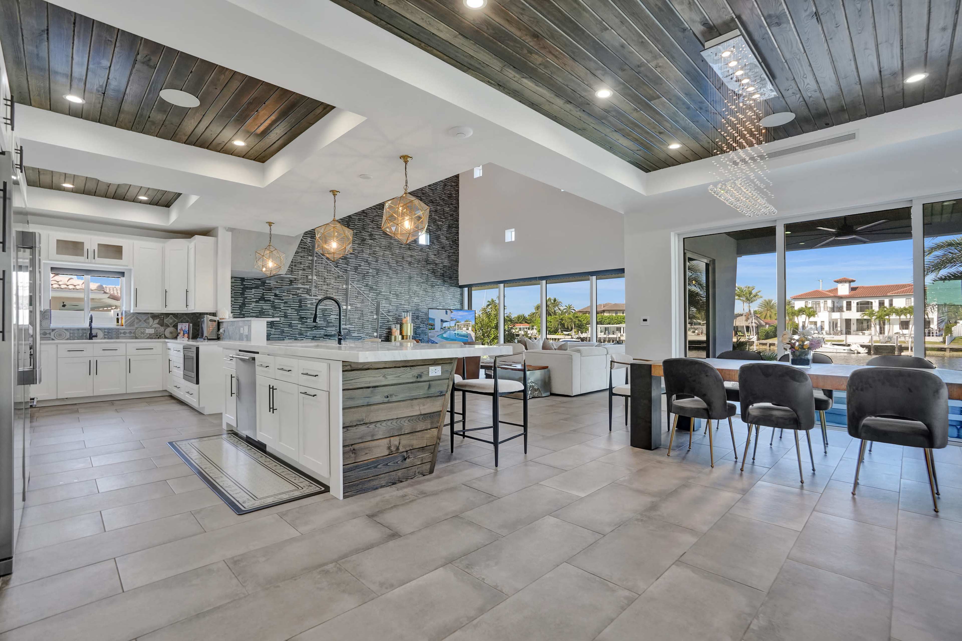The image shows a modern kitchen with white cabinets, a large island, and an adjoining dining area that features a scenic view through large windows.