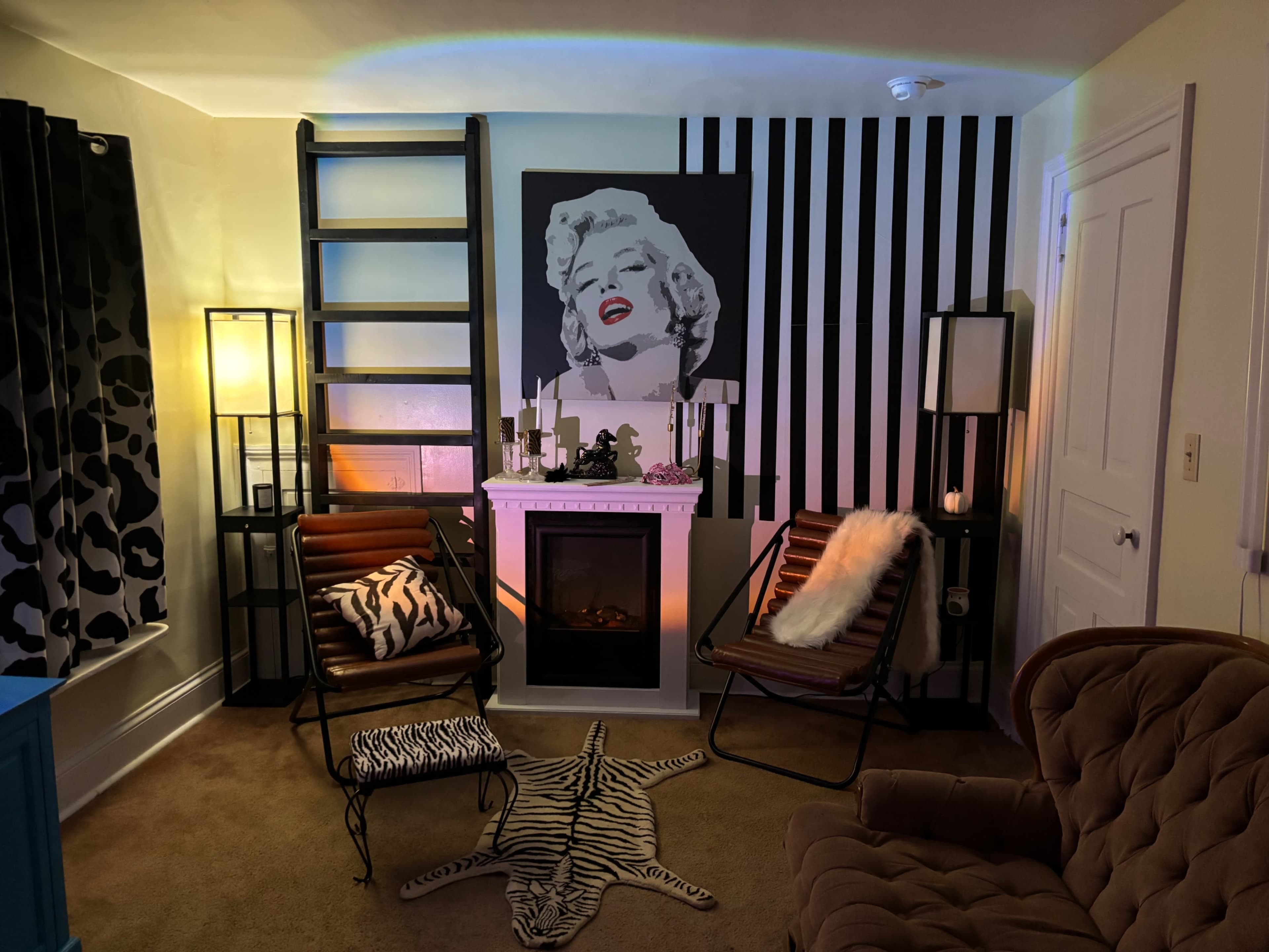 The image shows a living room corner featuring two striped chairs, a decorative ladder, a faux fireplace, and a large artwork of a woman on the wall.