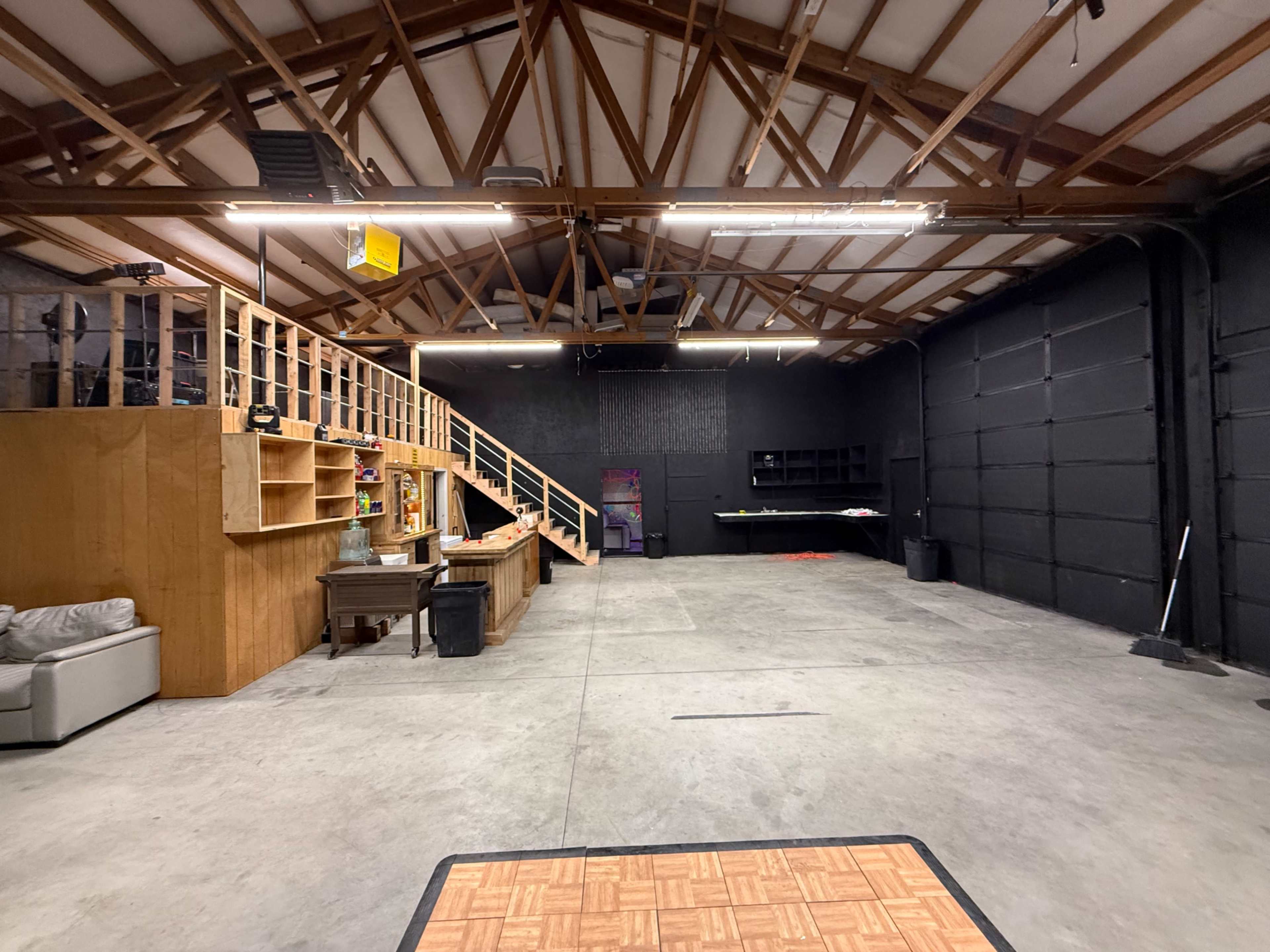 The image shows a spacious garage interior with wooden shelves, a staircase, and a large black wall.