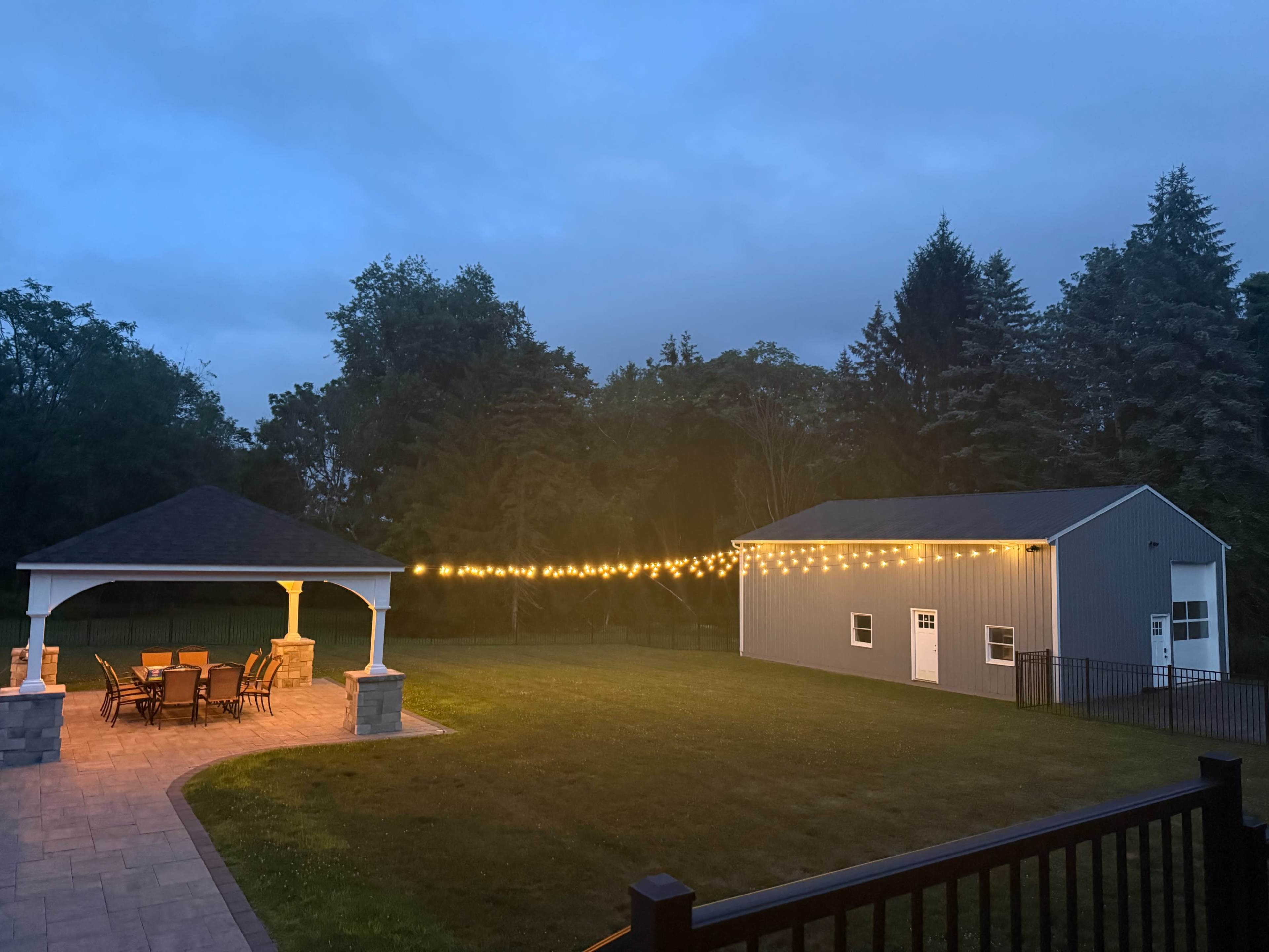 A gazebo with string lights illuminates a seating area in a yard next to a large gray shed under a twilight sky.