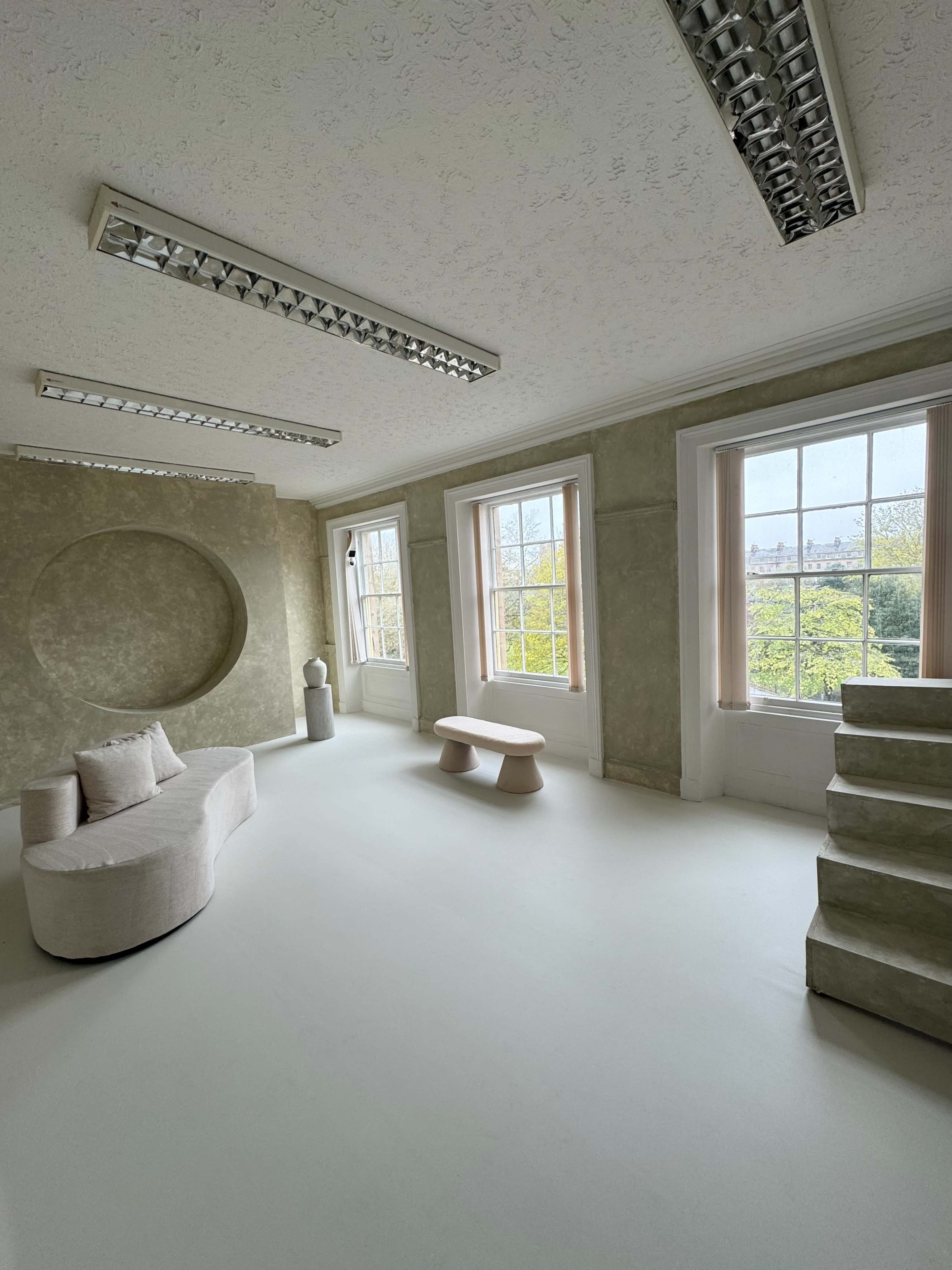 The image shows a spacious, minimalist room with large windows, a curved sofa, a low table, and a staircase.