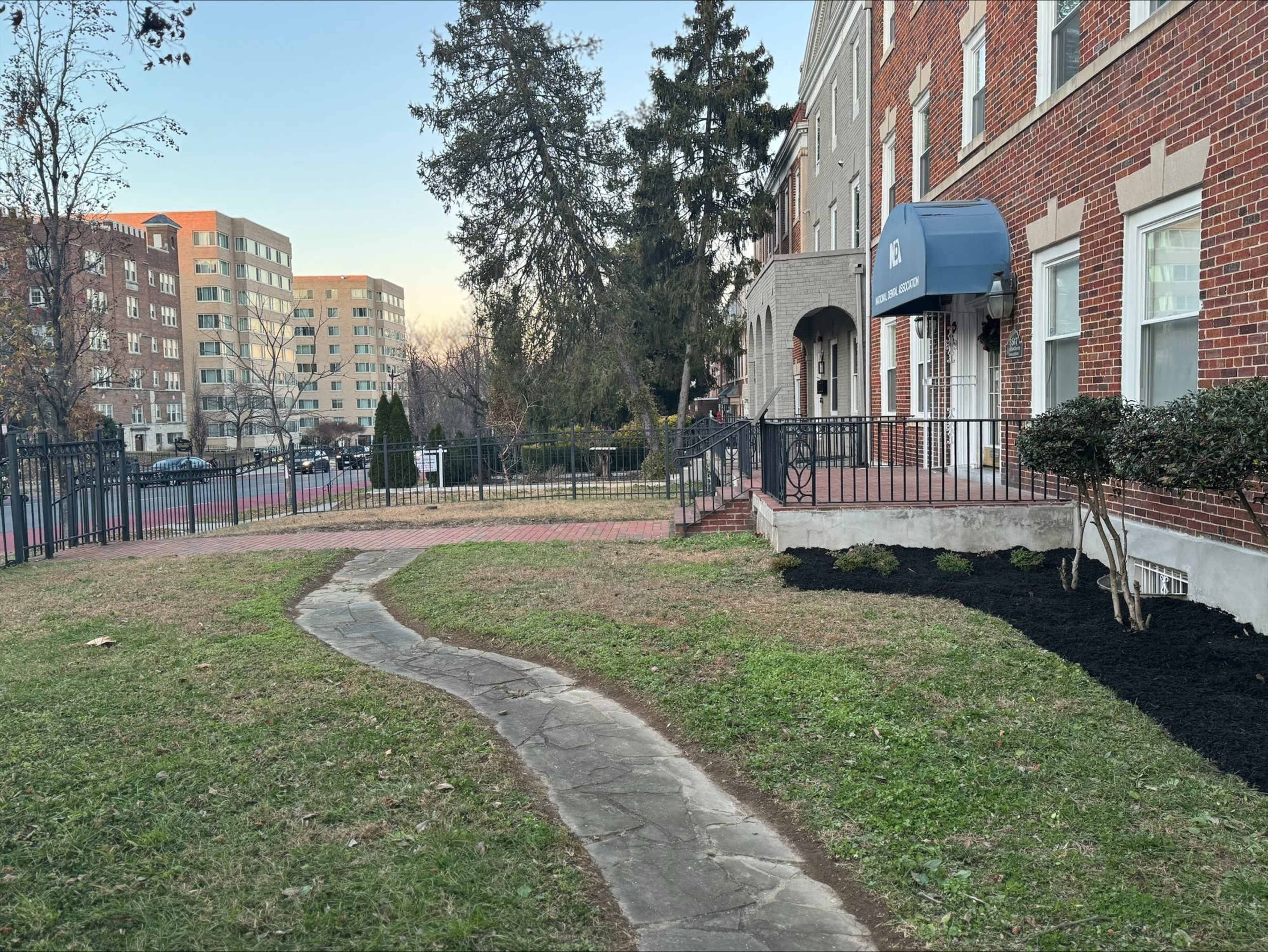A winding stone pathway leads from a brick building with an awning to a grassy area, with trees and other buildings visible in the background.