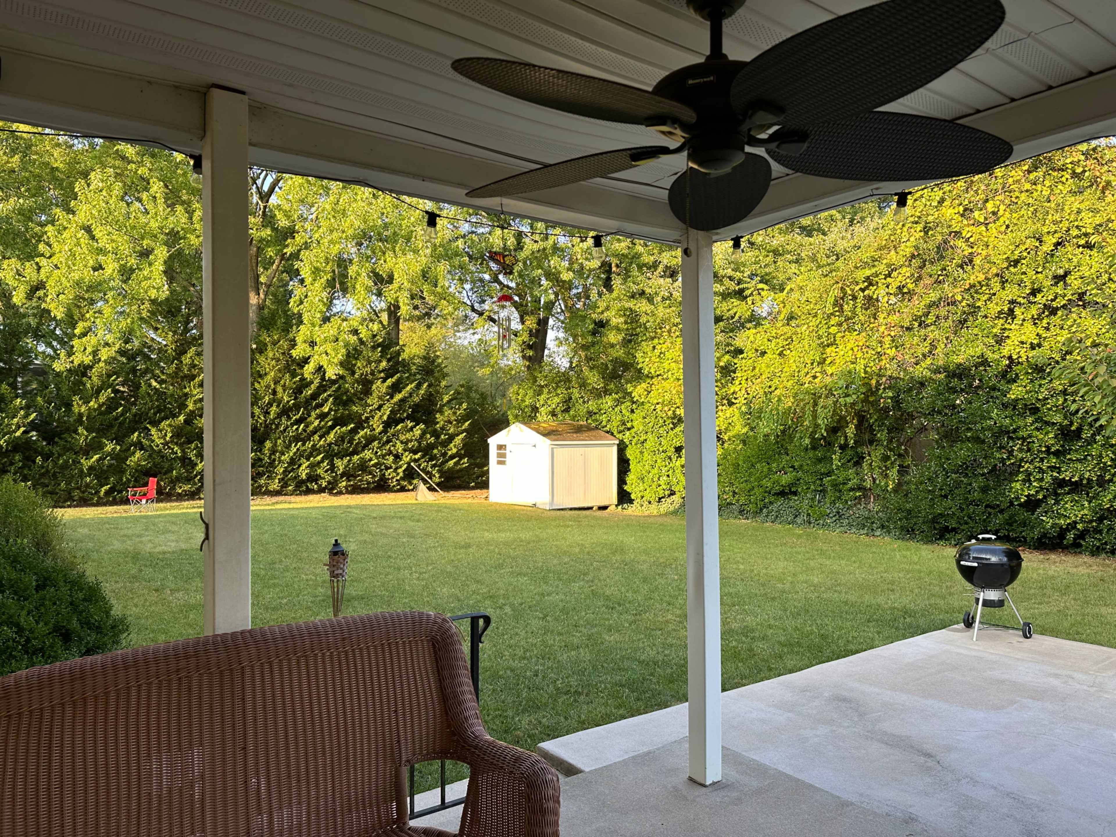 A wicker chair sits on a covered patio overlooking a grassy backyard with a storage shed and a charcoal grill.