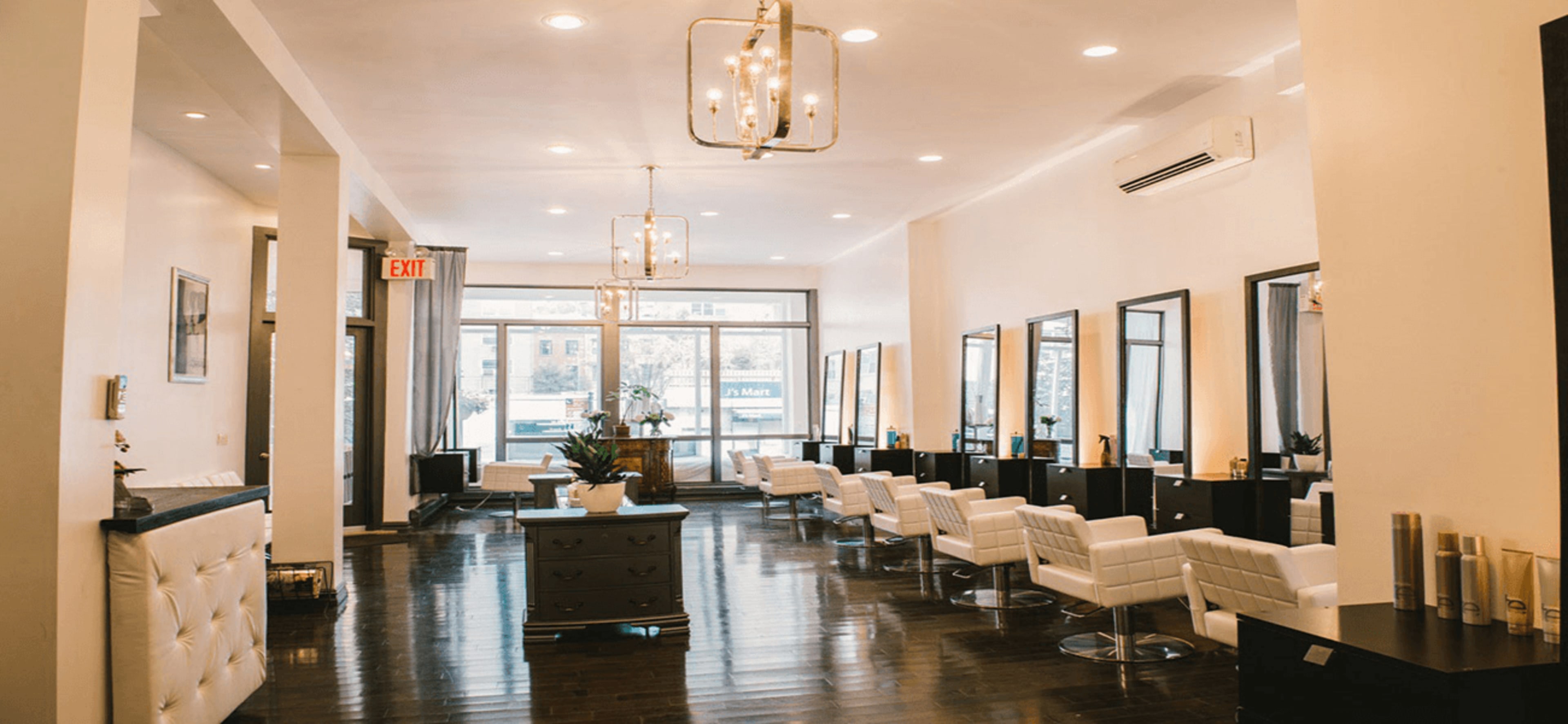 The image shows a modern hair salon with several white styling chairs arranged in front of large mirrors, illuminated by decorative light fixtures.