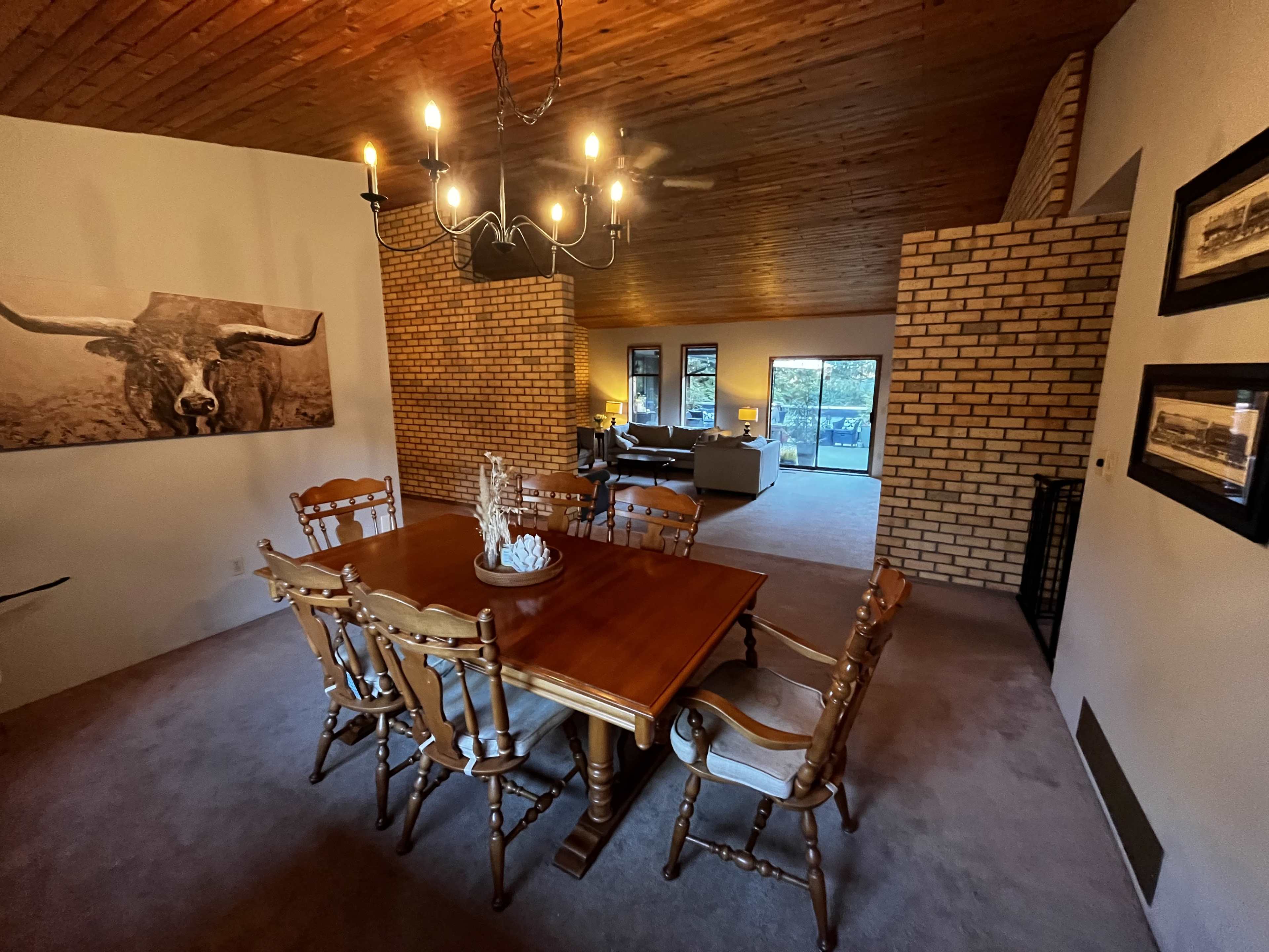 A dining area with a wooden table and chairs, adjacent to a living space visible through a doorway, featuring a brick wall and large windows.