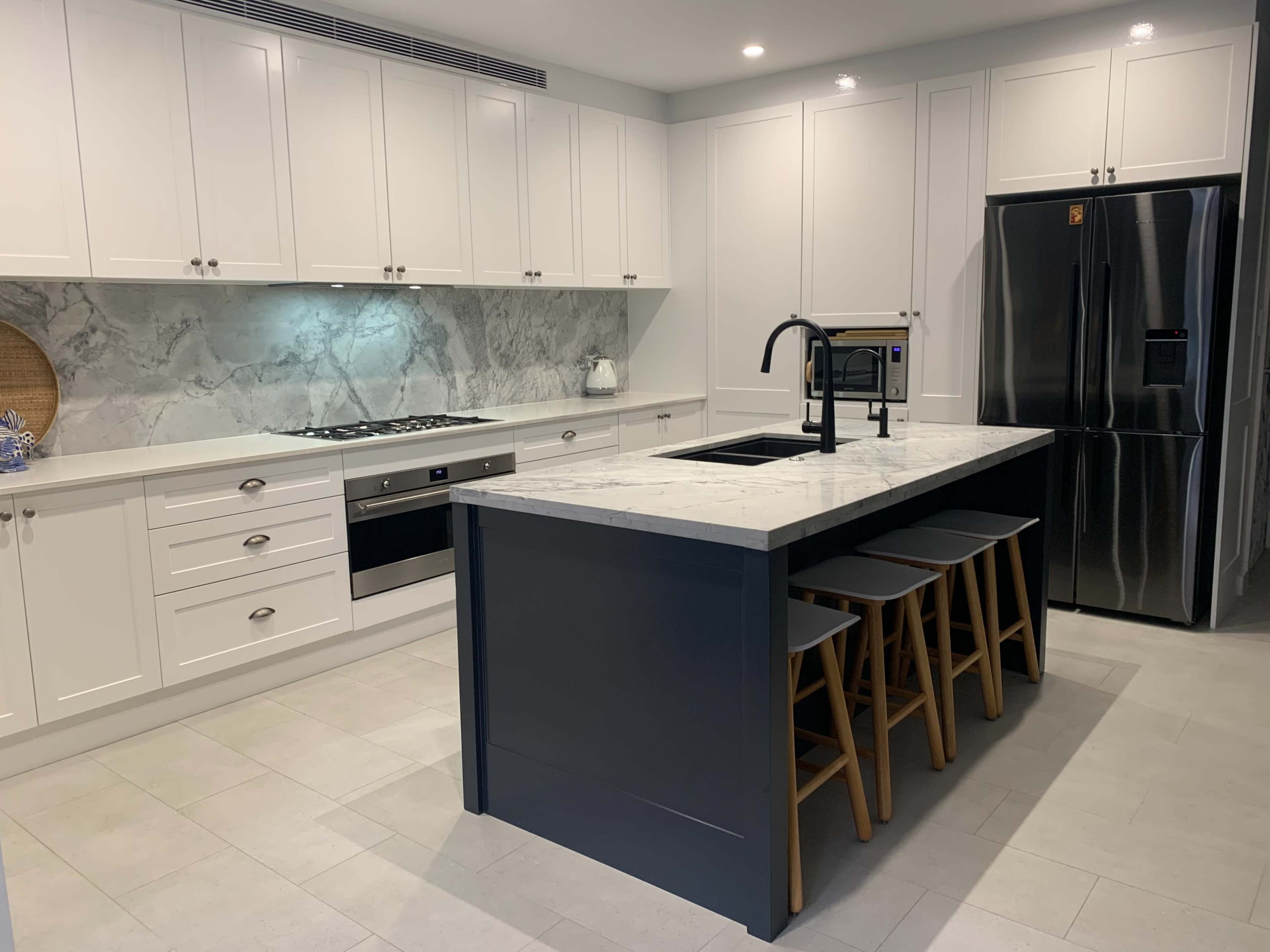 The image shows a modern kitchen featuring white cabinetry, a marble countertop island with three wooden stools, and stainless steel appliances.