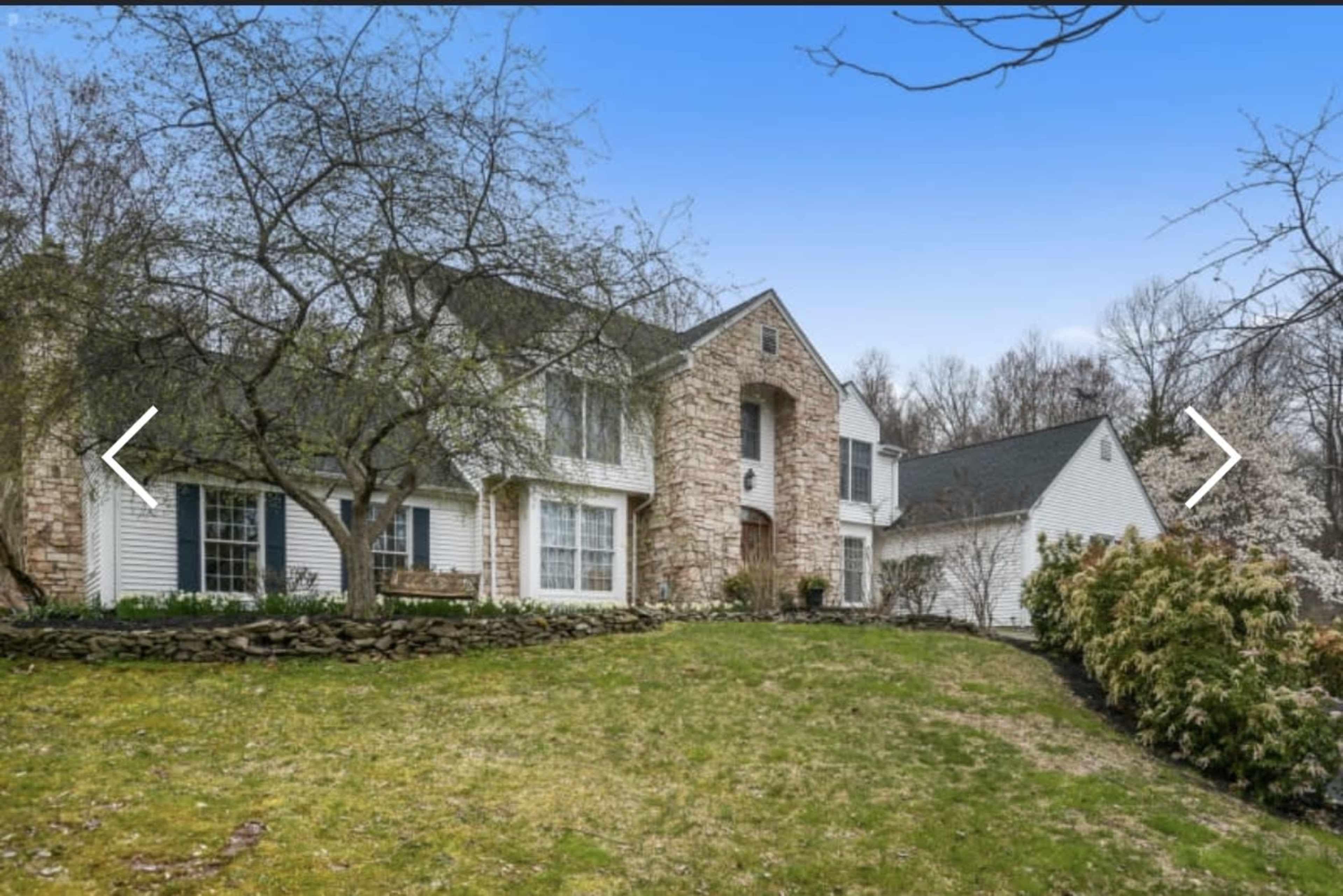 A two-story house with a stone facade and white siding sits on a landscaped lawn surrounded by trees and shrubs.