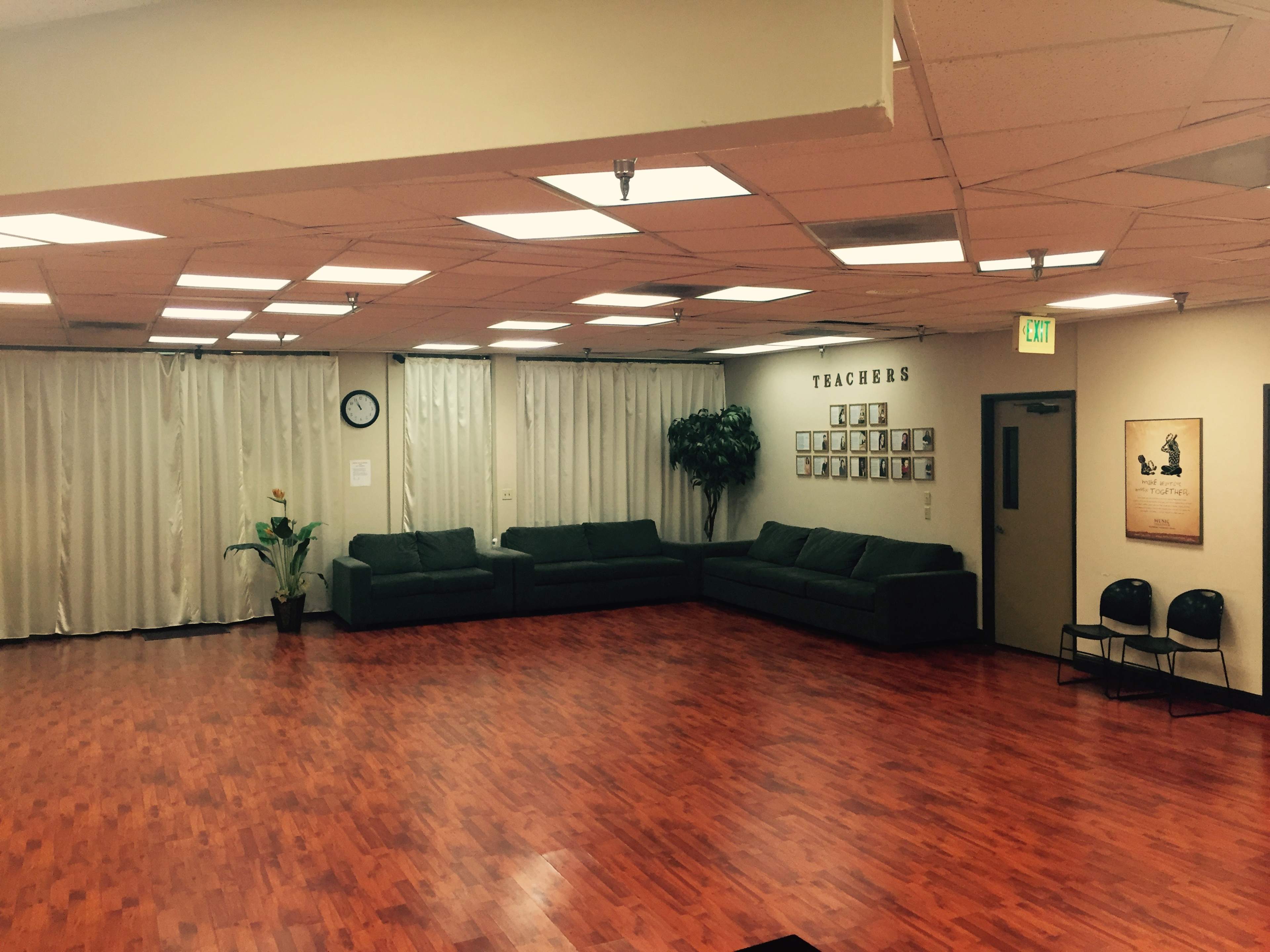 A spacious room with wooden flooring, green sofas arranged along the walls, and curtains covering the windows.