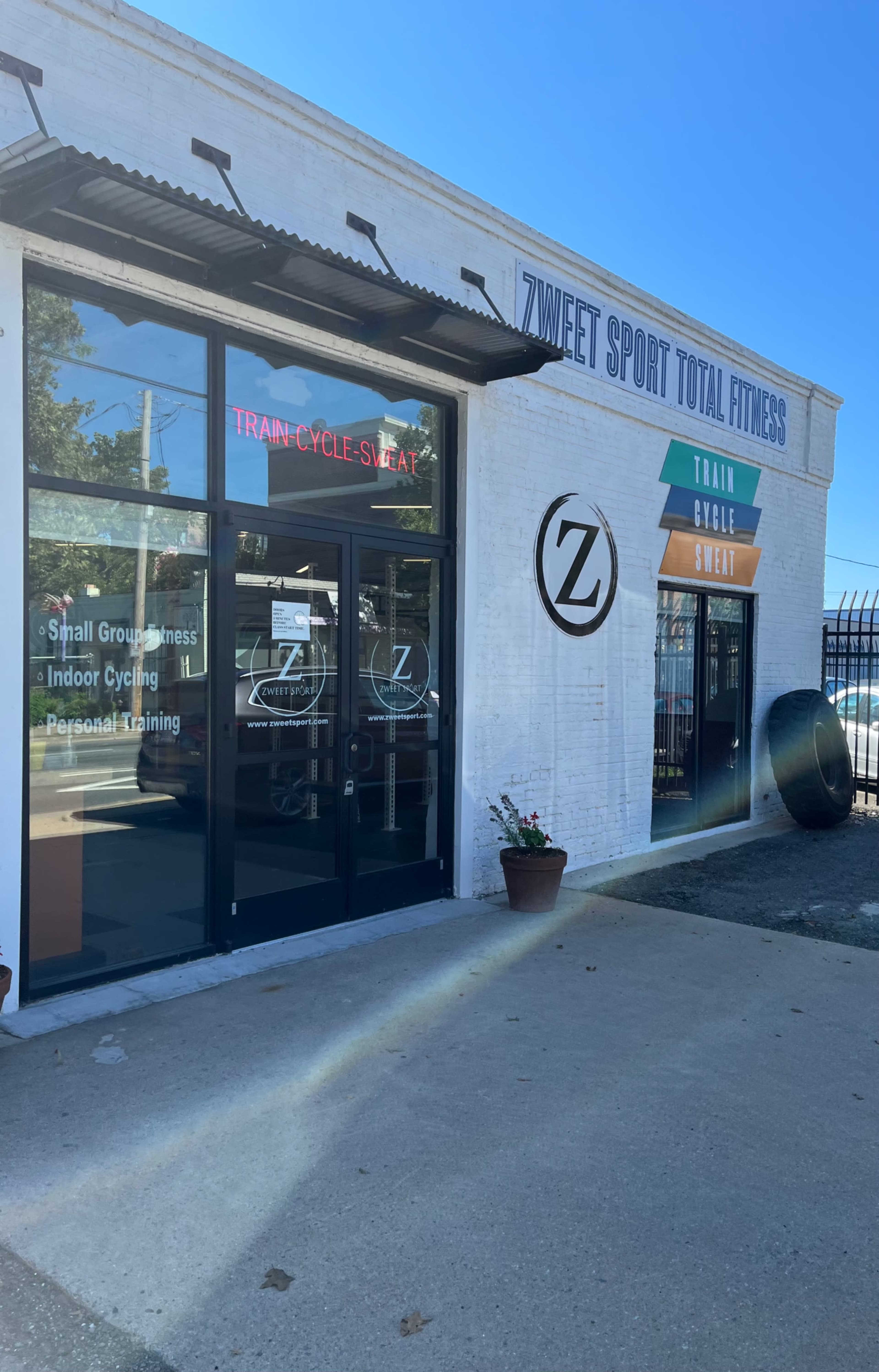 The image shows the exterior of a fitness studio named "Zweet Sport Total Fitness," featuring large glass windows and a sign displaying the services offered, including indoor cycling and personal training.