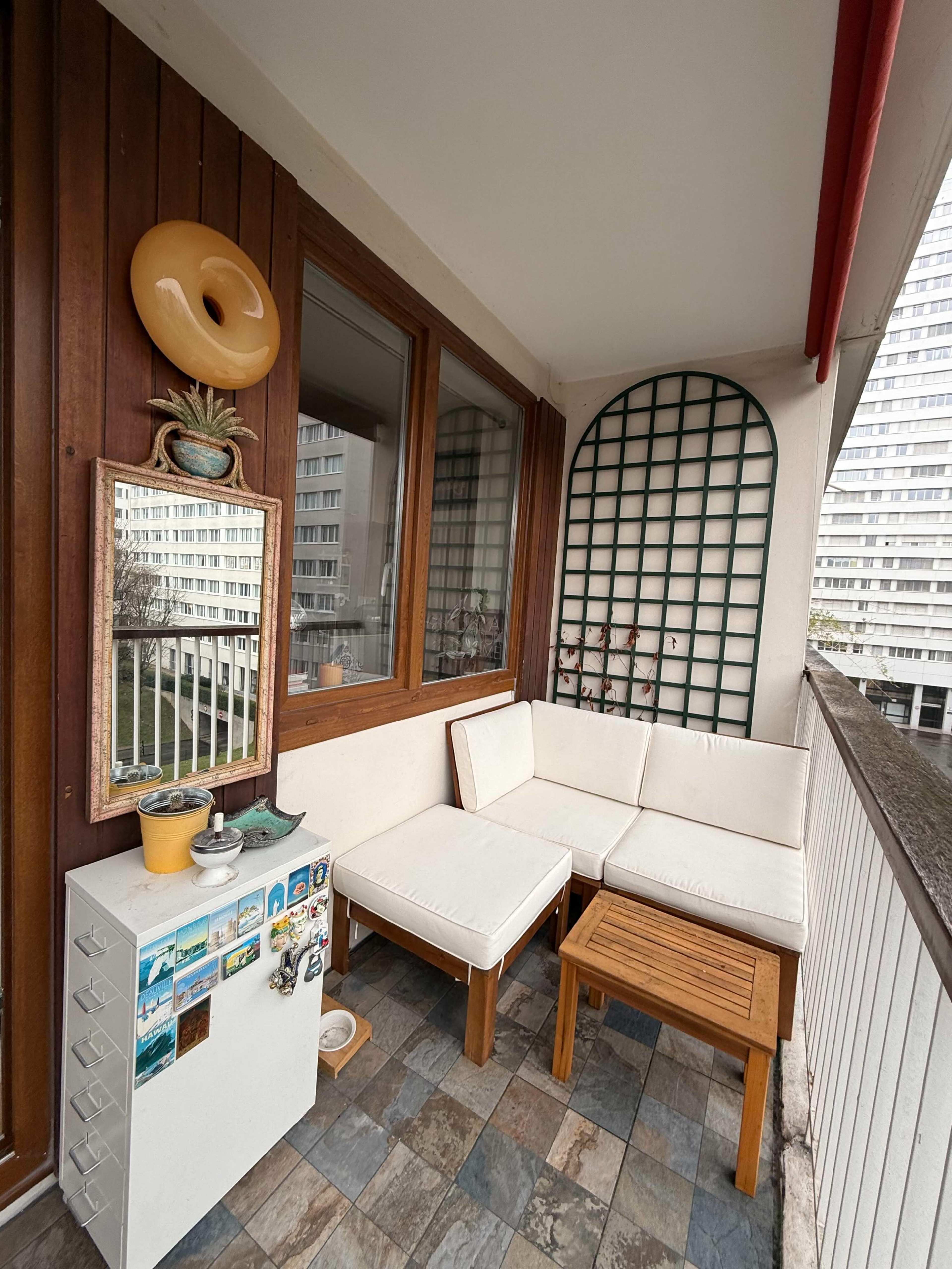 The image shows a small balcony area with a white couch, a wooden table, a cabinet, and a mirror, alongside decorative items against a backdrop of a residential building.