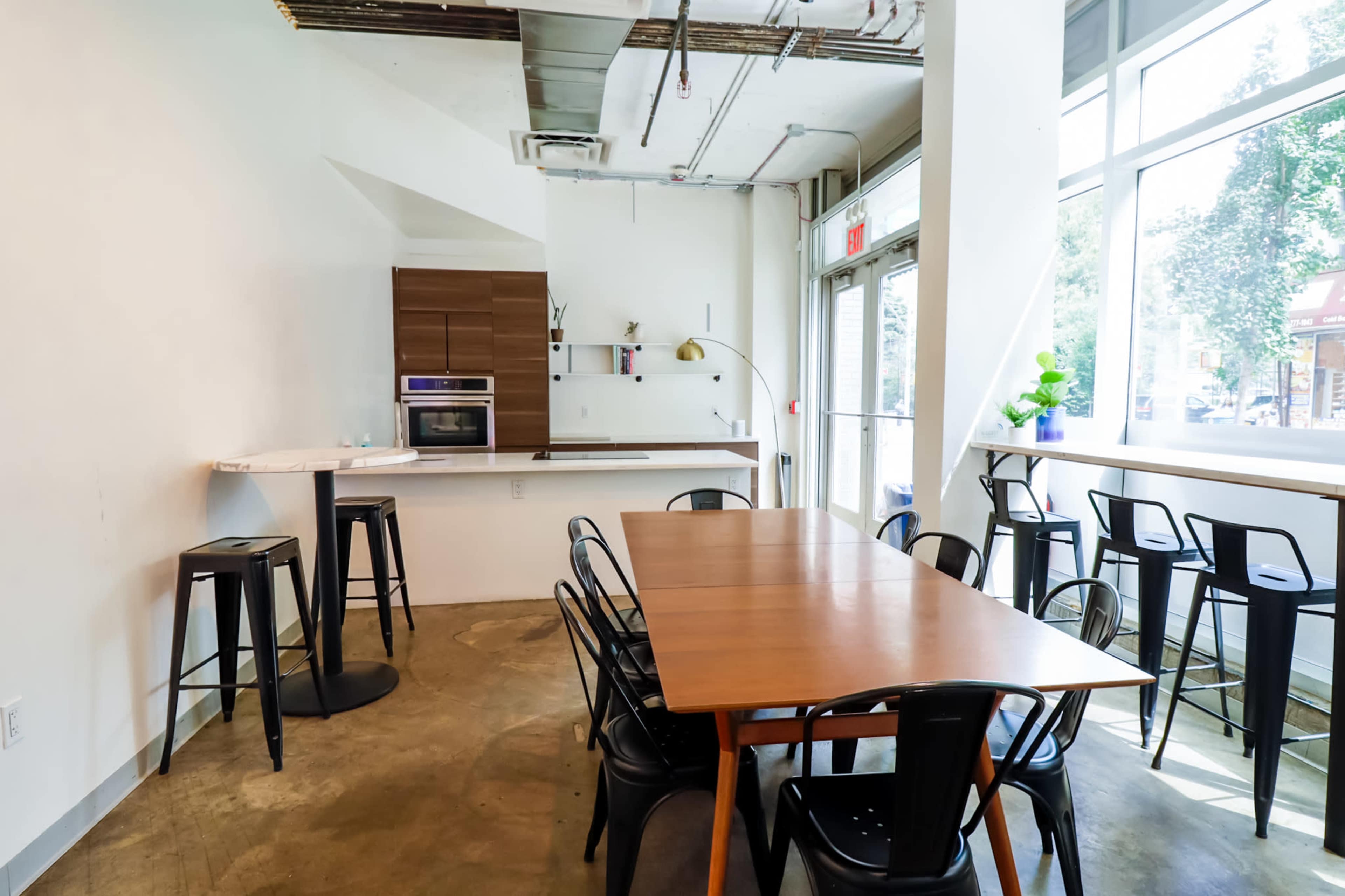 A modern, minimalist kitchen and dining area with a large wooden table surrounded by black chairs, set against a backdrop of large windows.