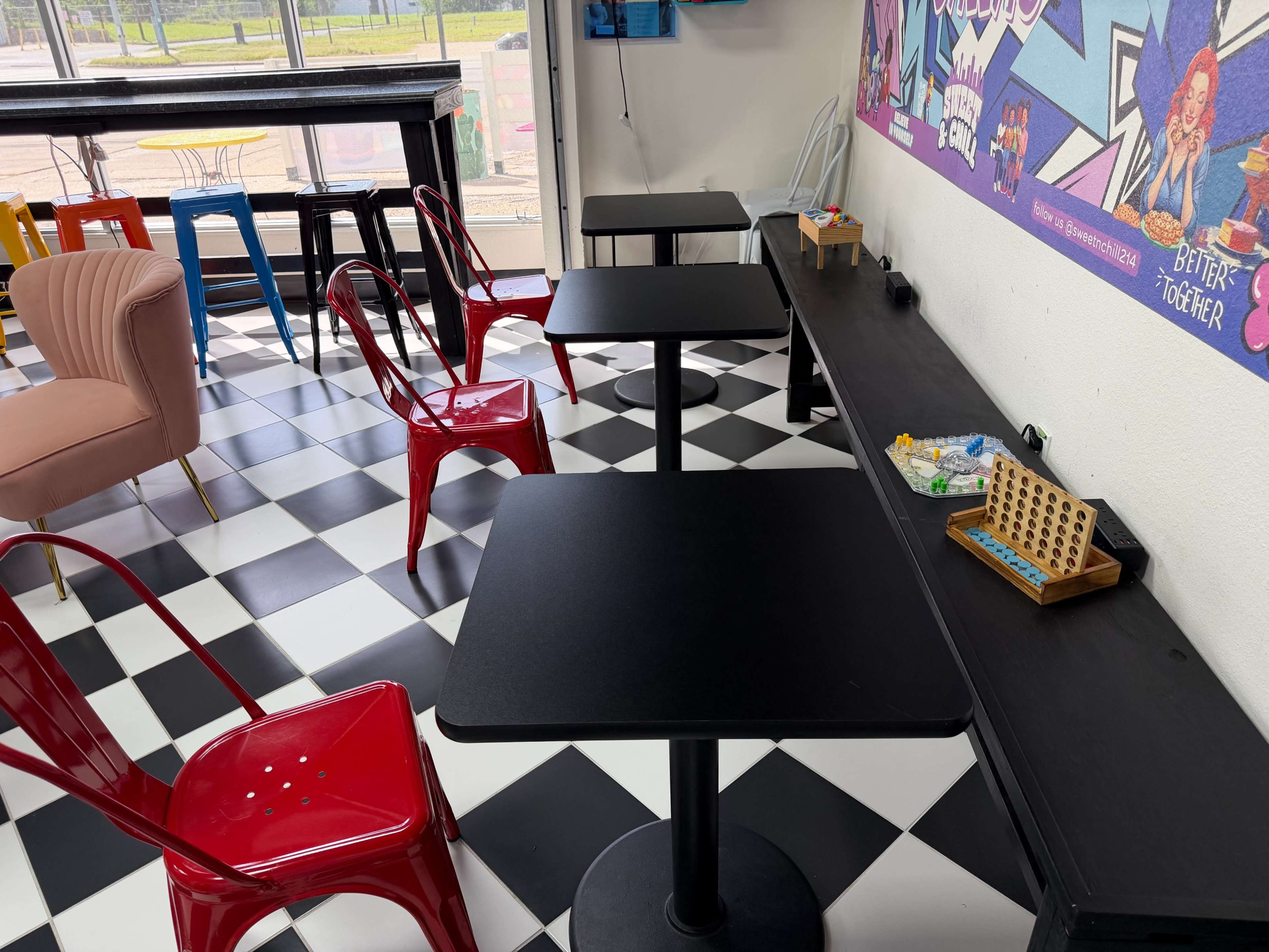 The image shows a modern café interior with black and white checkered floors, black tables, red and white chairs, and a colorful wall mural.