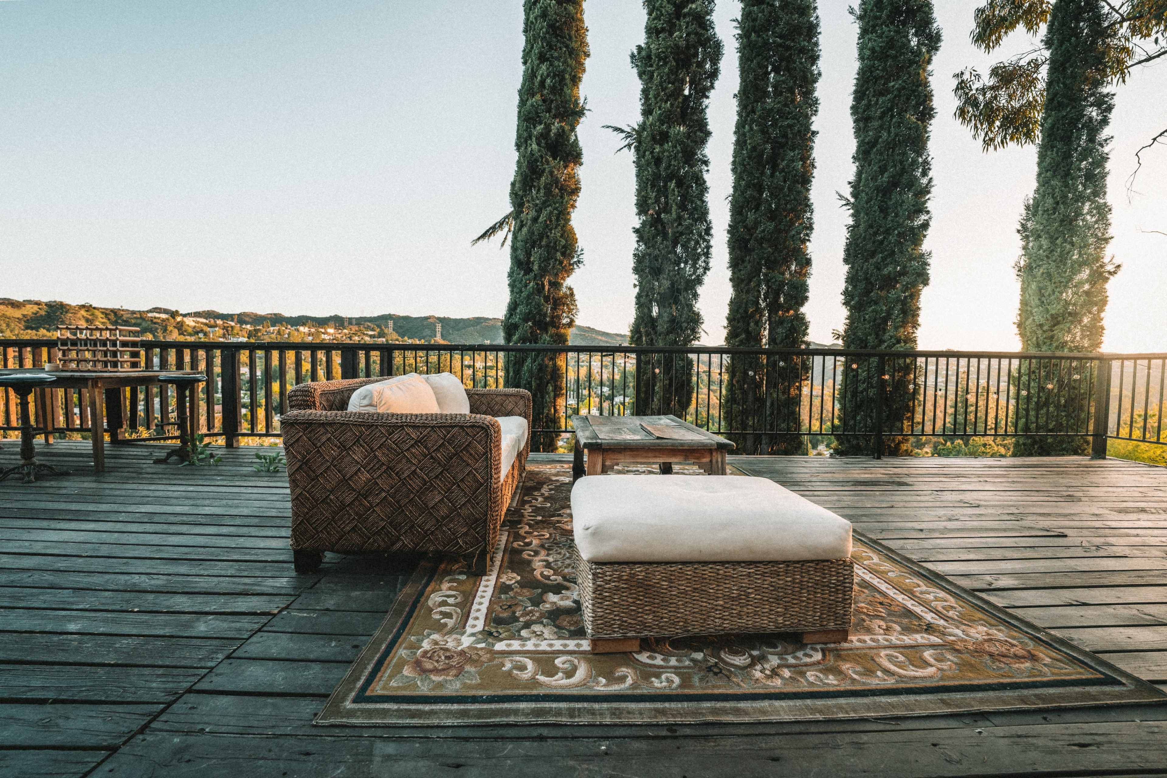 A wooden deck features a woven couch and an ottoman on a patterned rug, surrounded by tall trees and a scenic view.