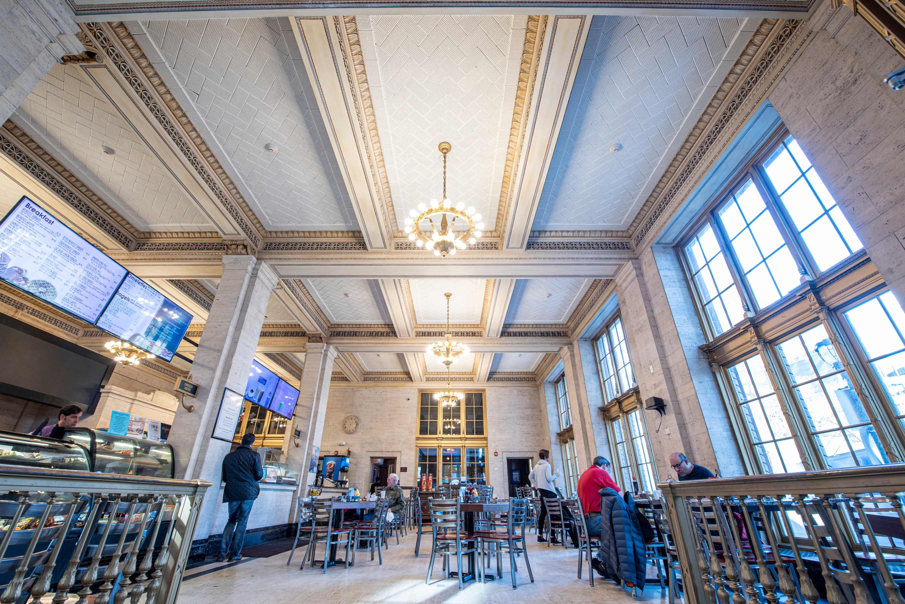 A spacious restaurant interior with high ceilings, large windows, and a mix of tables and chairs, occupied by a few patrons and staff.