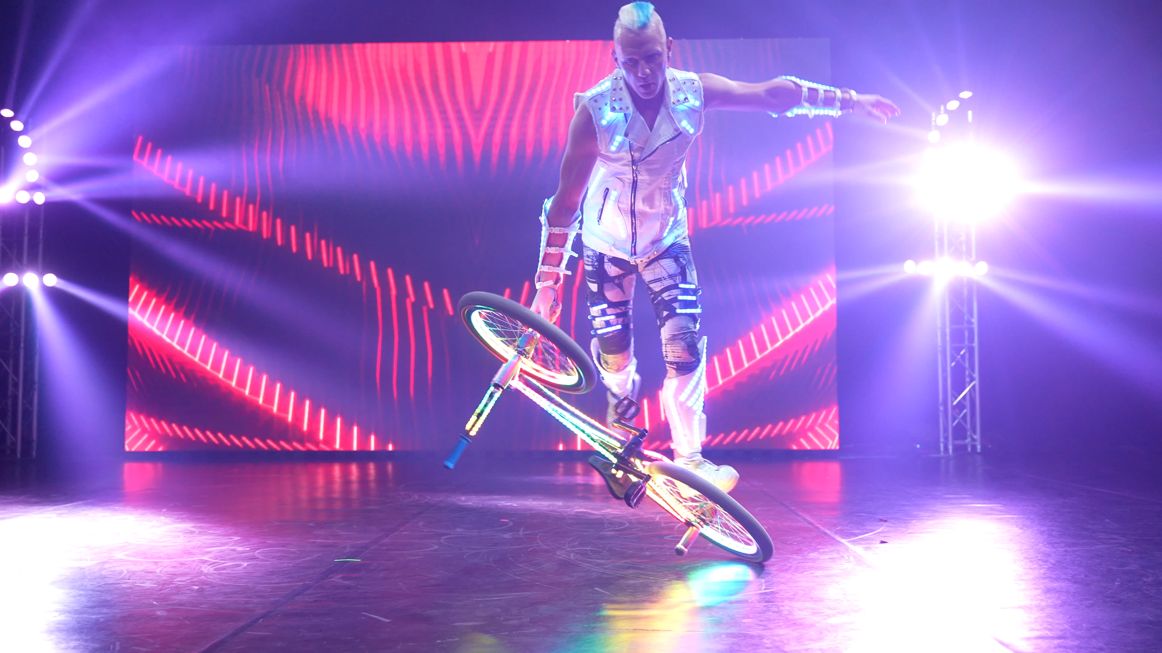 A performer executes a stunt on a bicycle under bright stage lights with a colorful digital backdrop.
