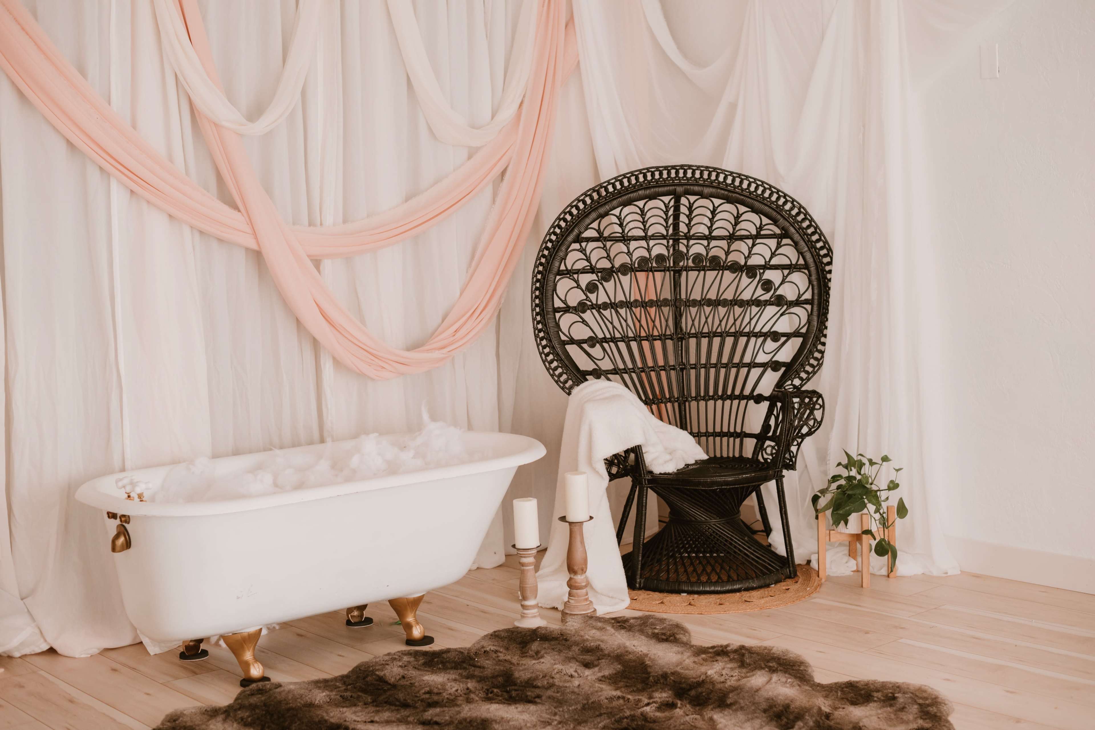 A vintage-style bathtub filled with bubbles sits next to a large, intricate wicker chair, accompanied by candles and a potted plant against a backdrop of draped fabric.