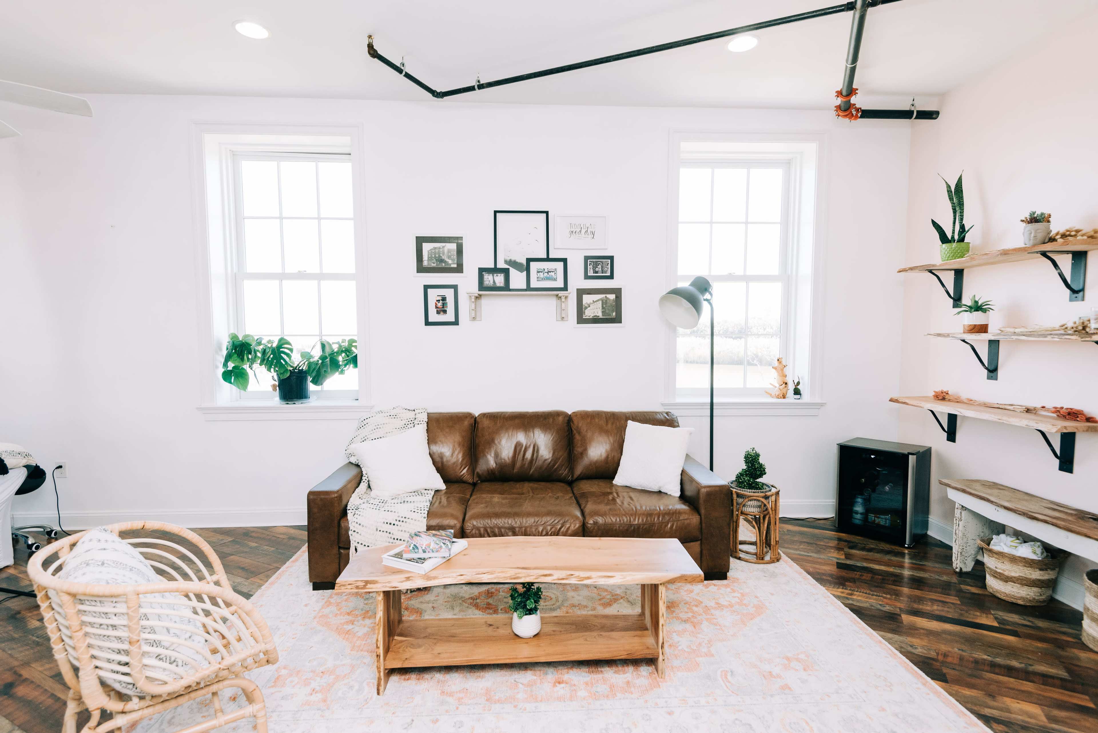 A living room features a brown leather sofa, a wooden coffee table on a light rug, and plants on shelves and windowsills.