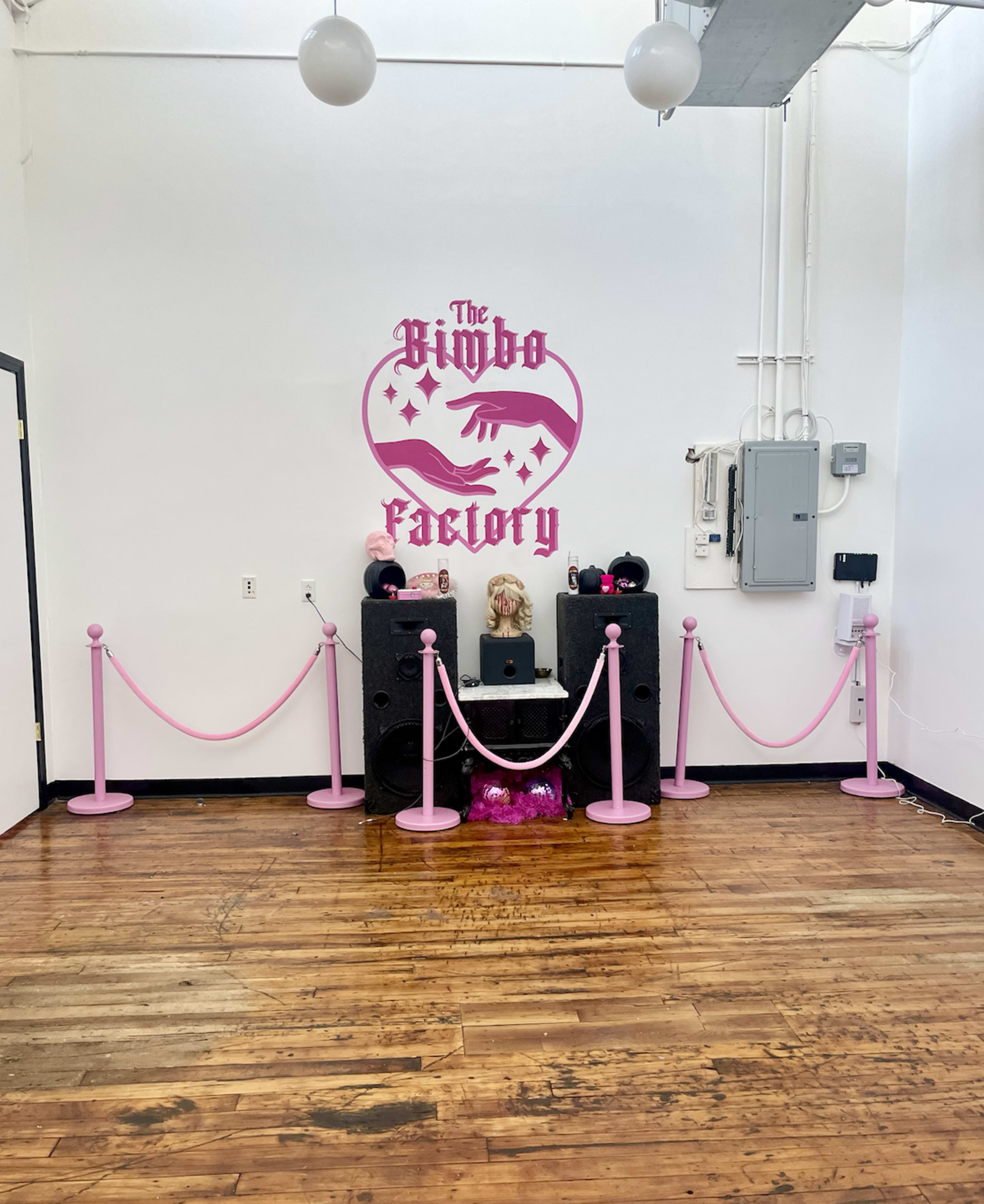 The image shows a pink-themed space called "The Bimbo Factory," featuring a decorative wall with a hand graphic and audio equipment flanked by pink ropes and stanchions.