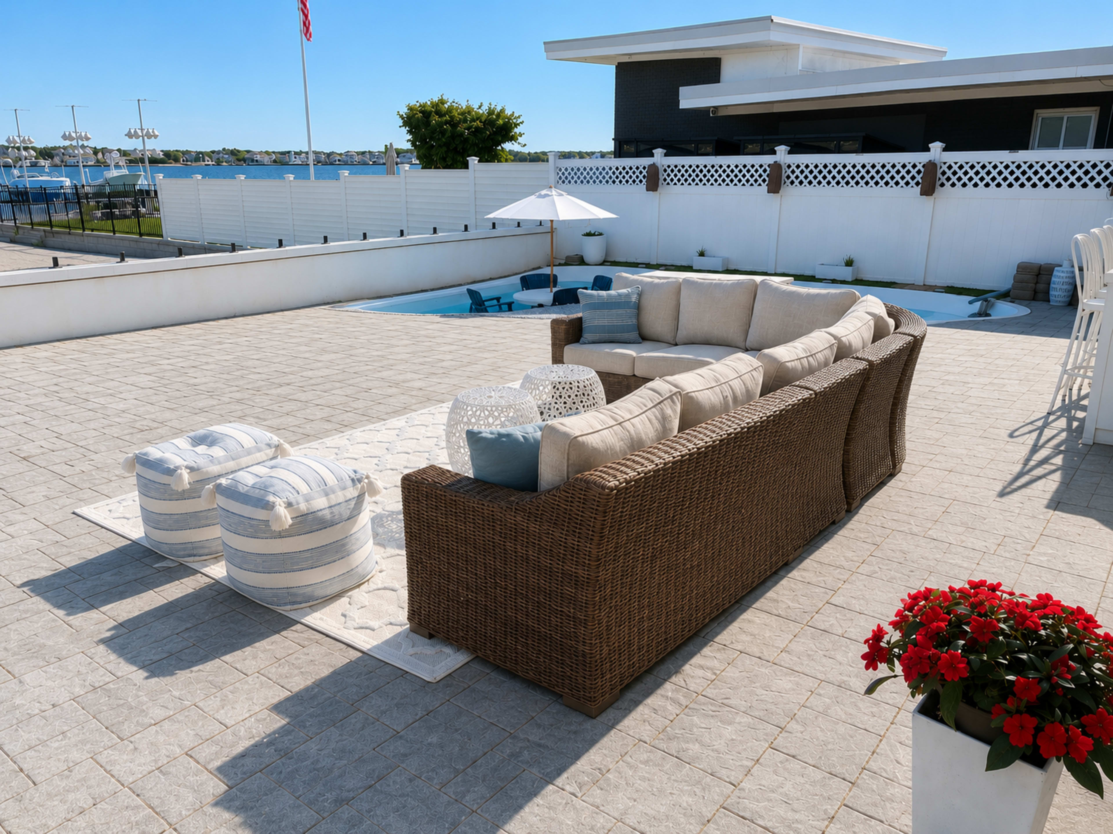 The image shows a patio area with a large sectional sofa, striped ottomans, a pool in the background, and a flower planter with red blooms.