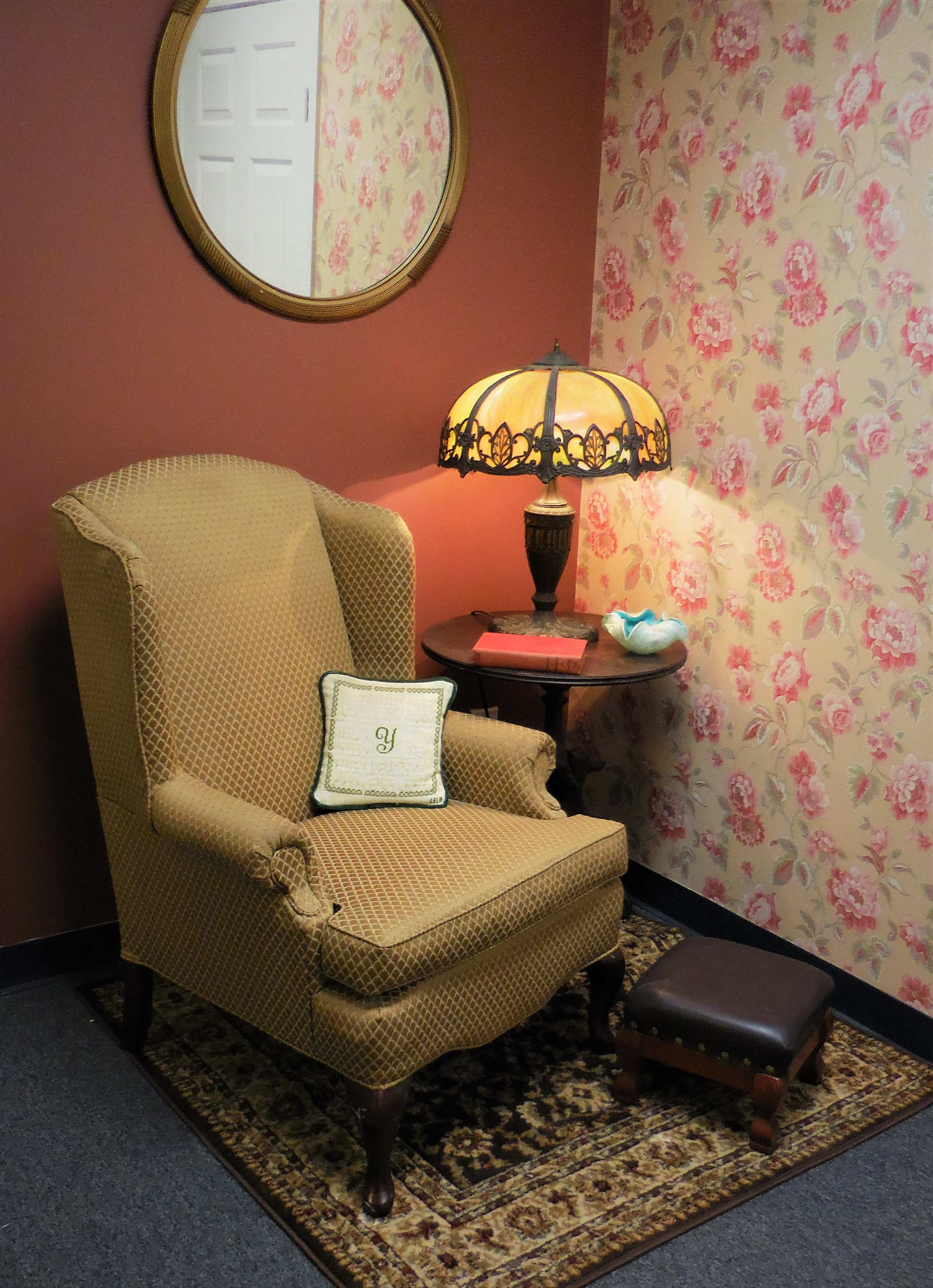 A cozy corner features a wingback chair with a decorative pillow, a round mirror on the wall, a lamp on a side table, and floral wallpaper.