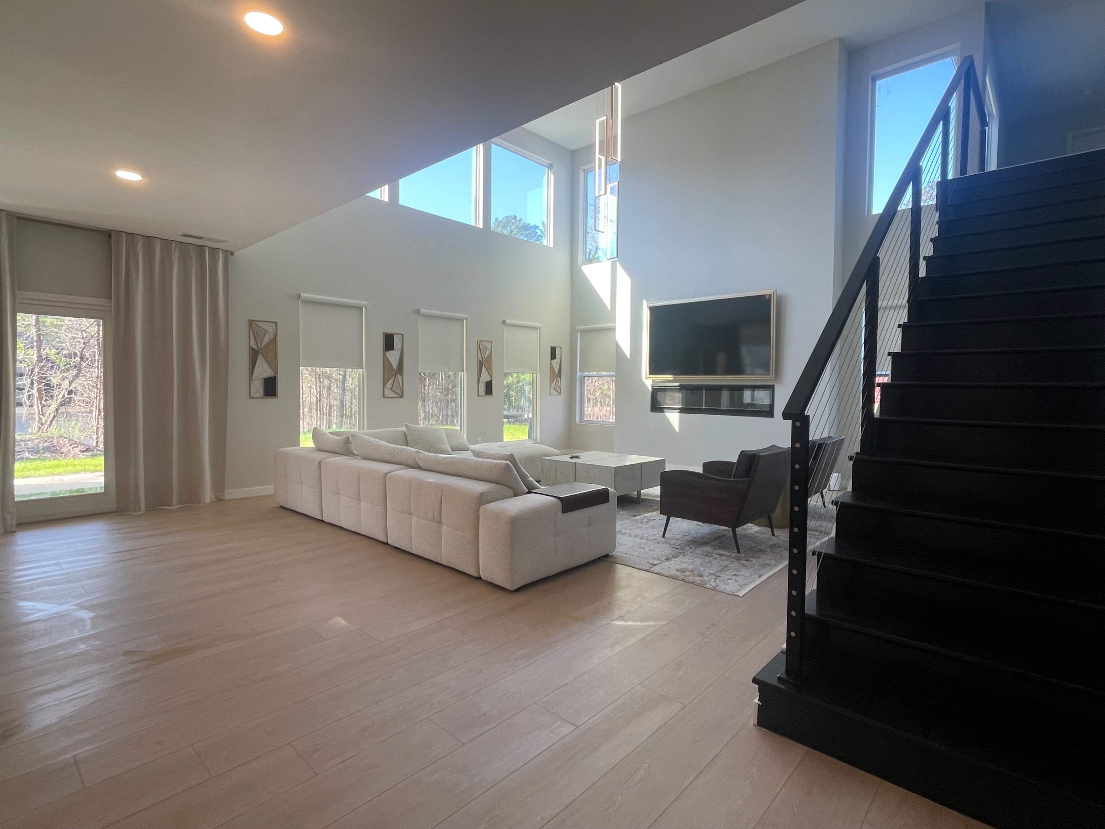 A modern living room with a large sectional sofa, a coffee table, a mounted TV, and a staircase in a well-lit space featuring large windows.