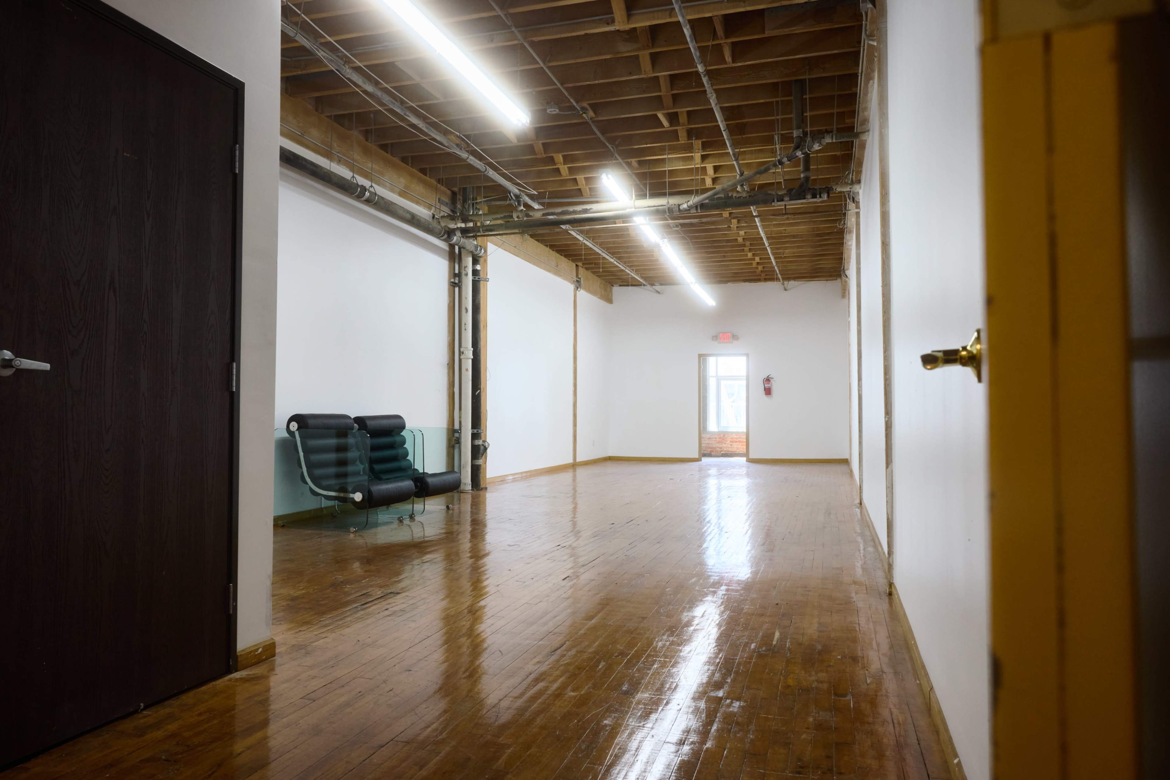 The image shows a spacious, empty room with polished wooden floors, exposed ceiling beams, and a green sofa along one wall.