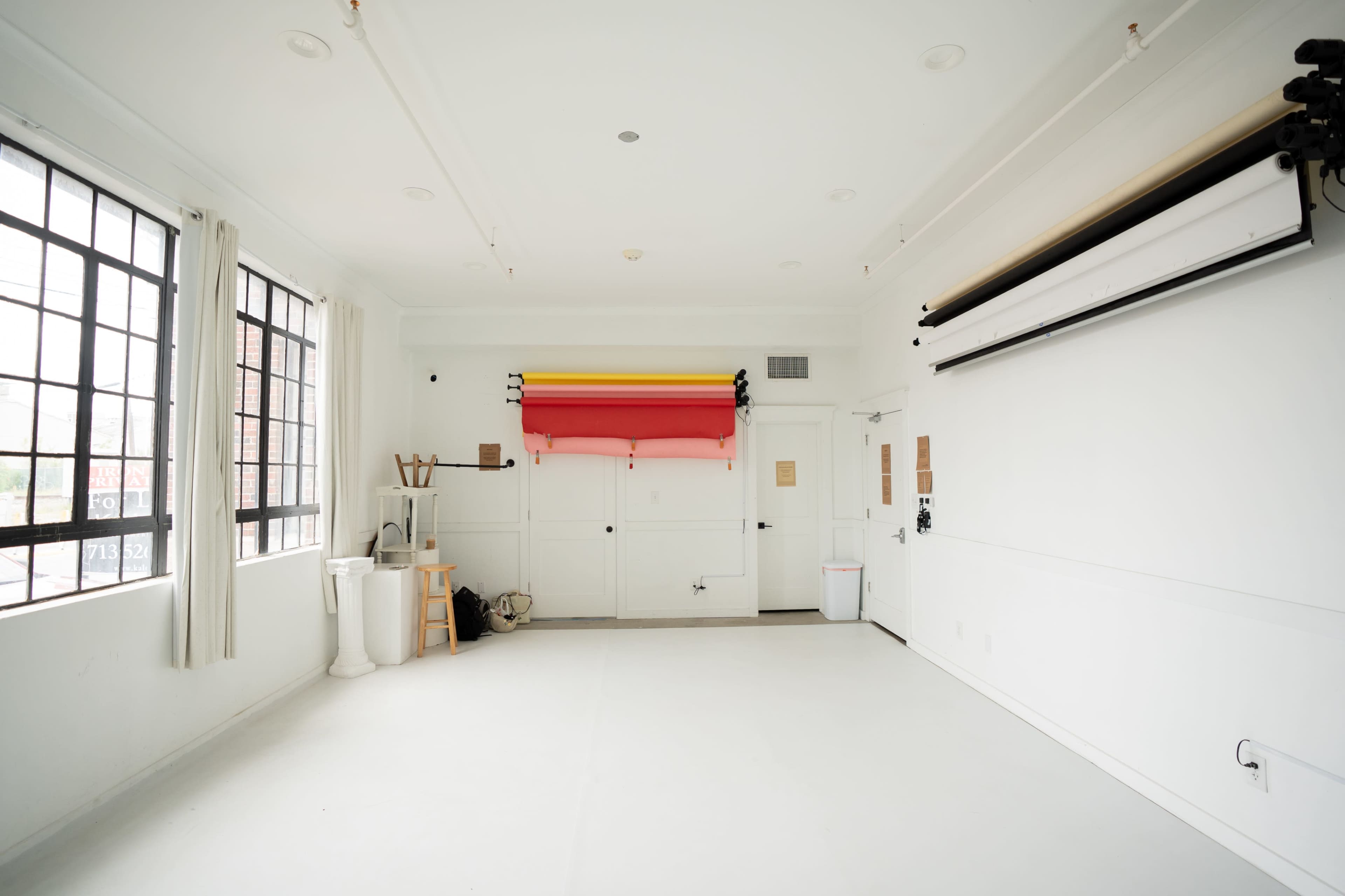 The image shows a minimalist photo studio with white walls, large windows, and a backdrop system featuring rolls of colored paper.