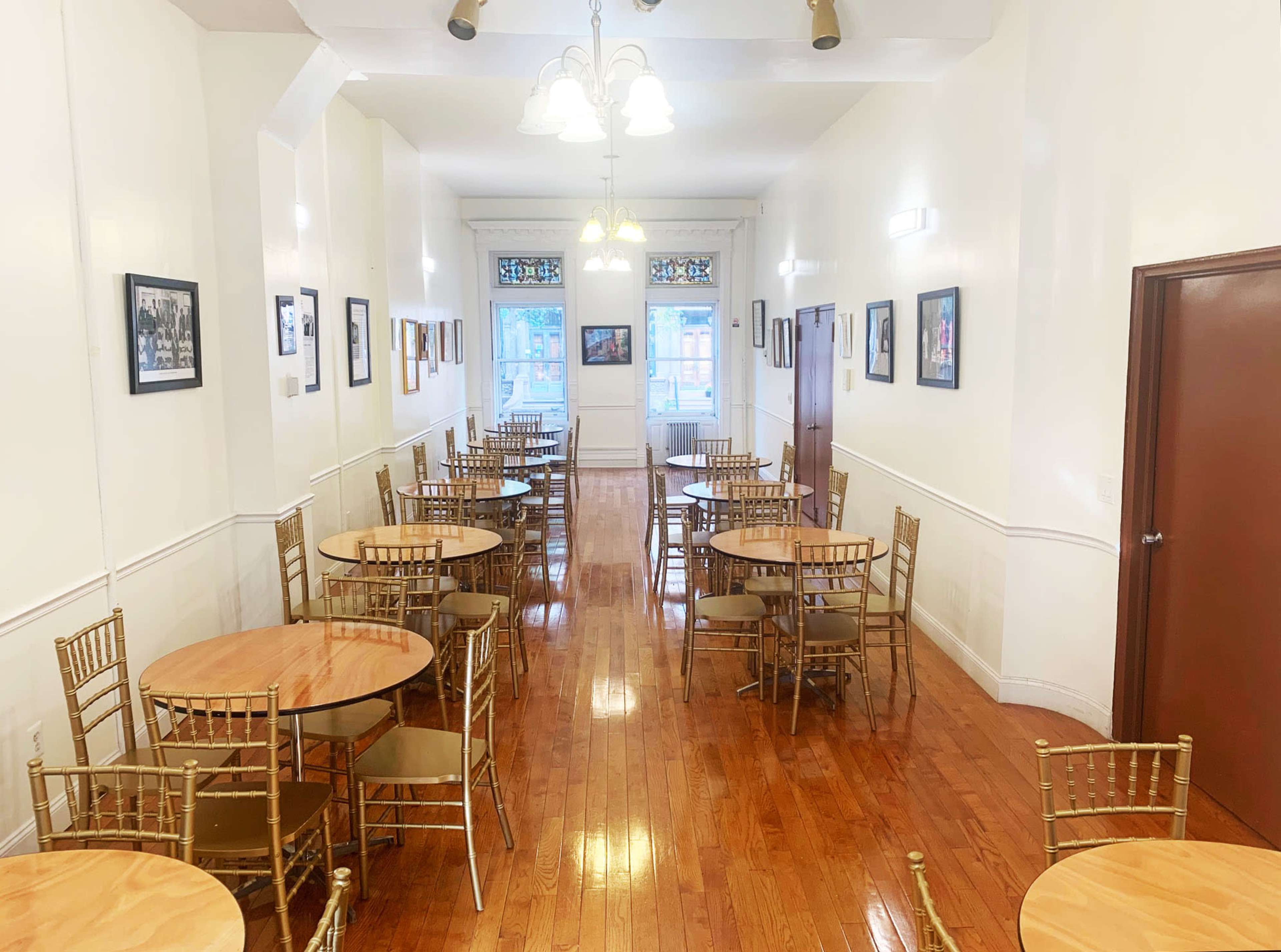 A long, narrow room features golden chairs and round wooden tables arranged neatly, with framed photographs on the white walls.