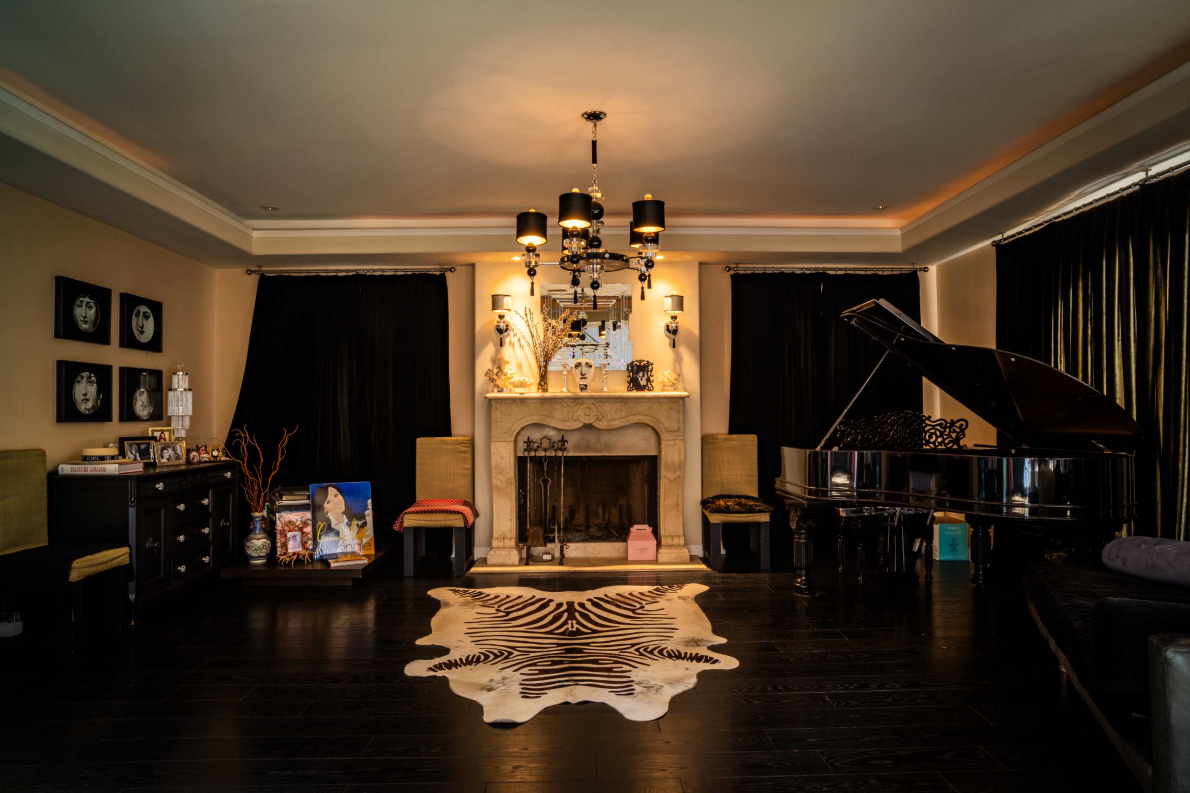 The living room features a grand piano beside a stone fireplace, adorned with decorative items and framed artwork, complemented by dark curtains and an animal print rug on a black wooden floor.