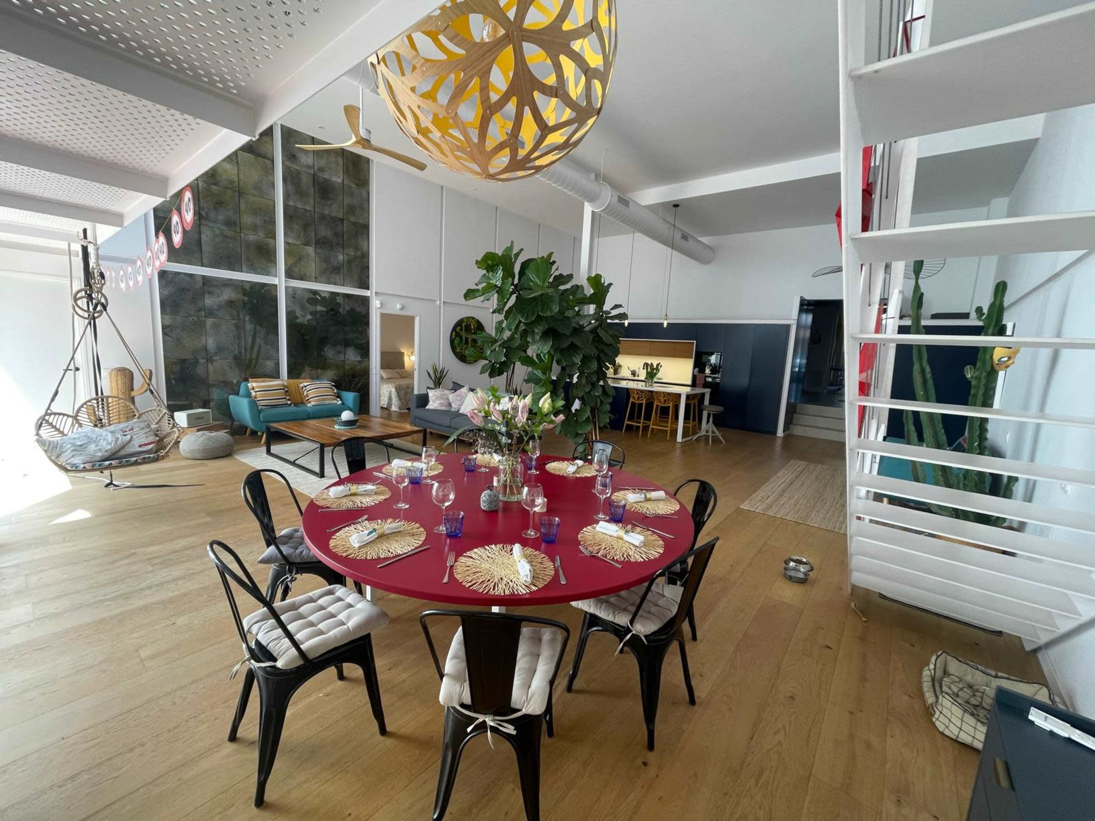 A large round table with a red tabletop is set for a meal in a bright, open-concept living space featuring modern furniture and decorative plants.