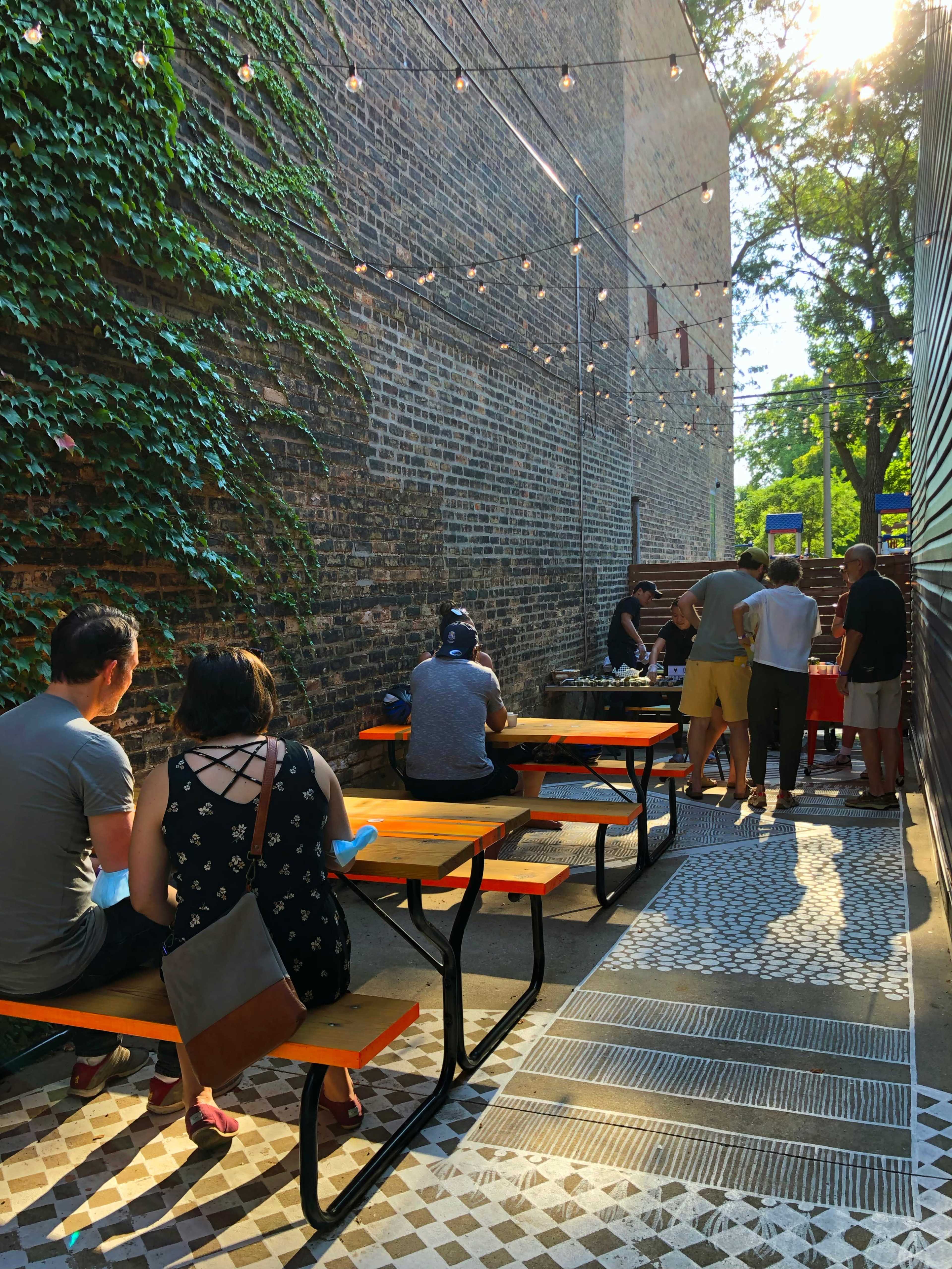 A narrow outdoor space with wooden picnic tables, string lights overhead, and people gathered along a brick wall covered in greenery.