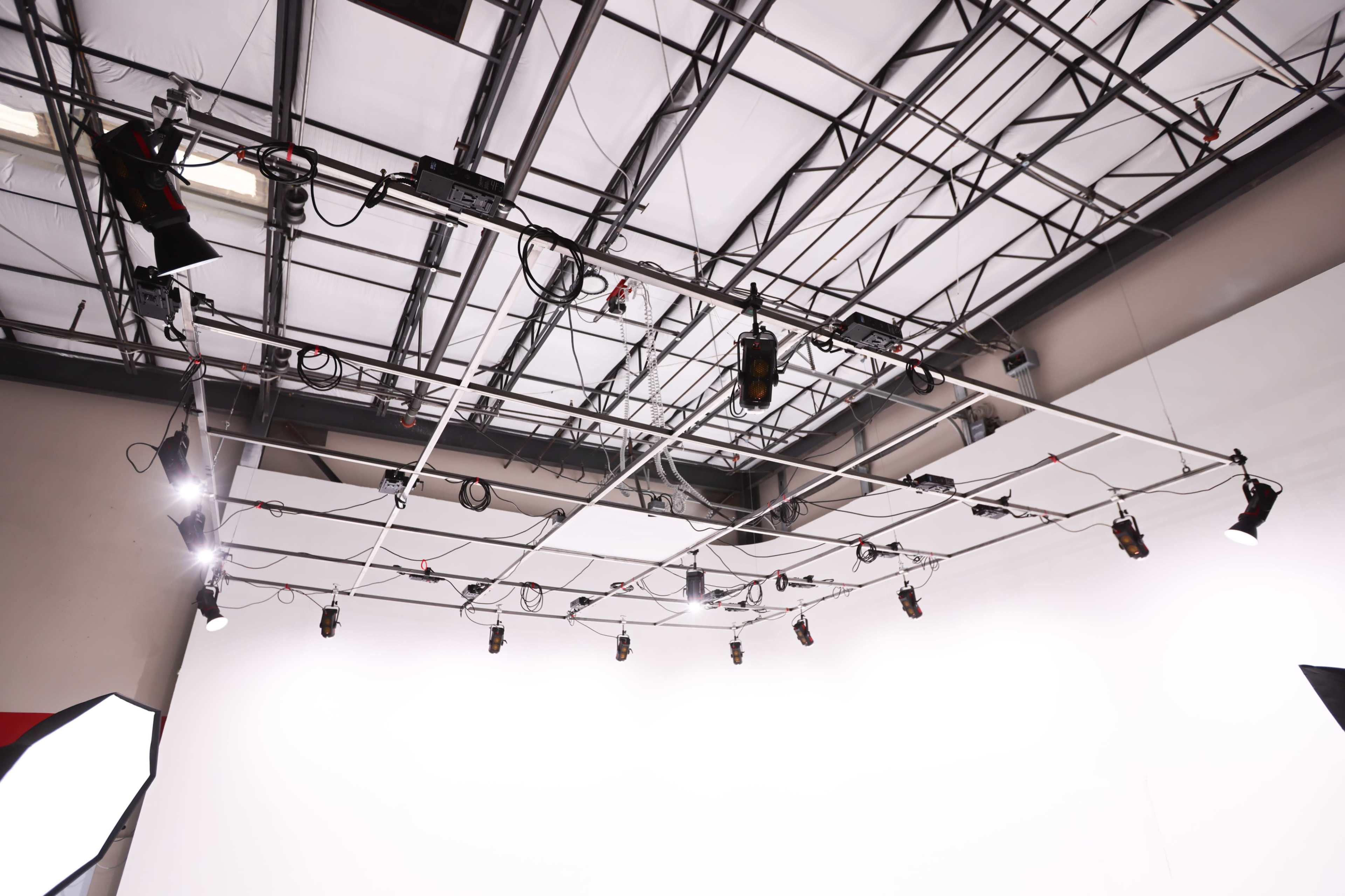 A ceiling grid with multiple lights and equipment suspended above a blank white backdrop in a studio space.