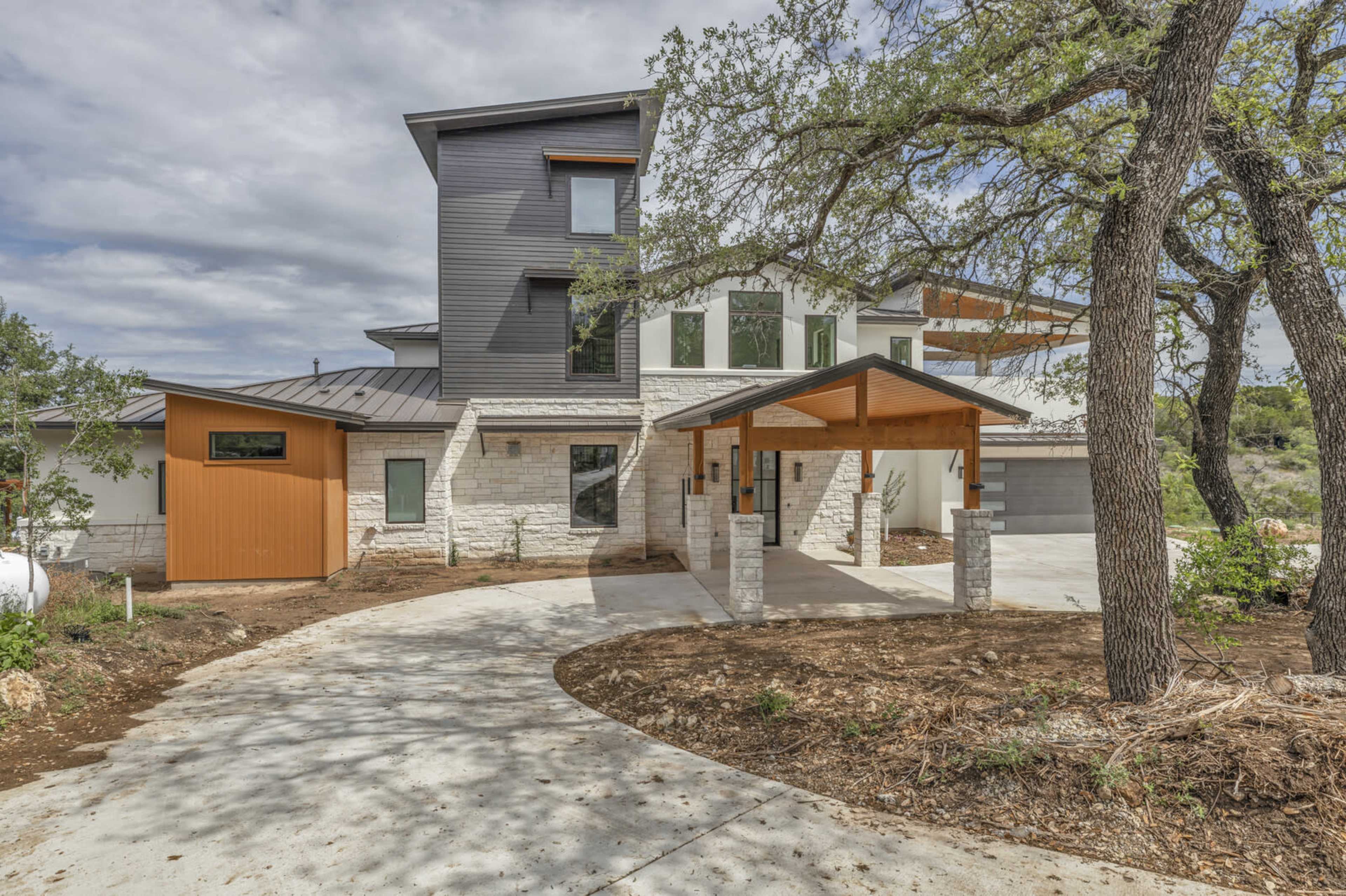 A modern, multi-story house with a combination of stone and wood elements, a circular driveway, and several trees surrounding the property.