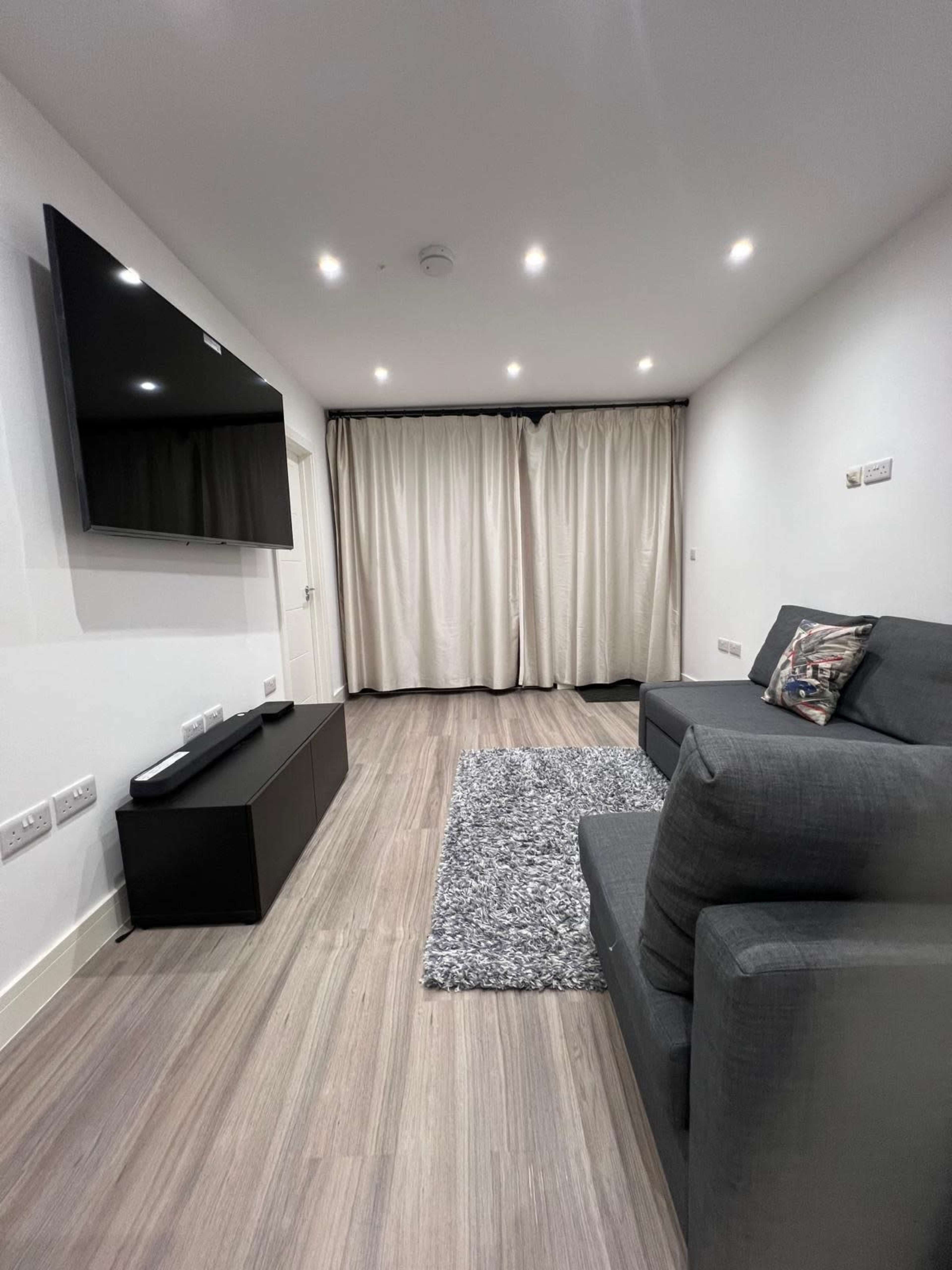 The image shows a modern living room with a gray couch, a black media console with a television mounted on the wall, and light-colored curtains covering a window.