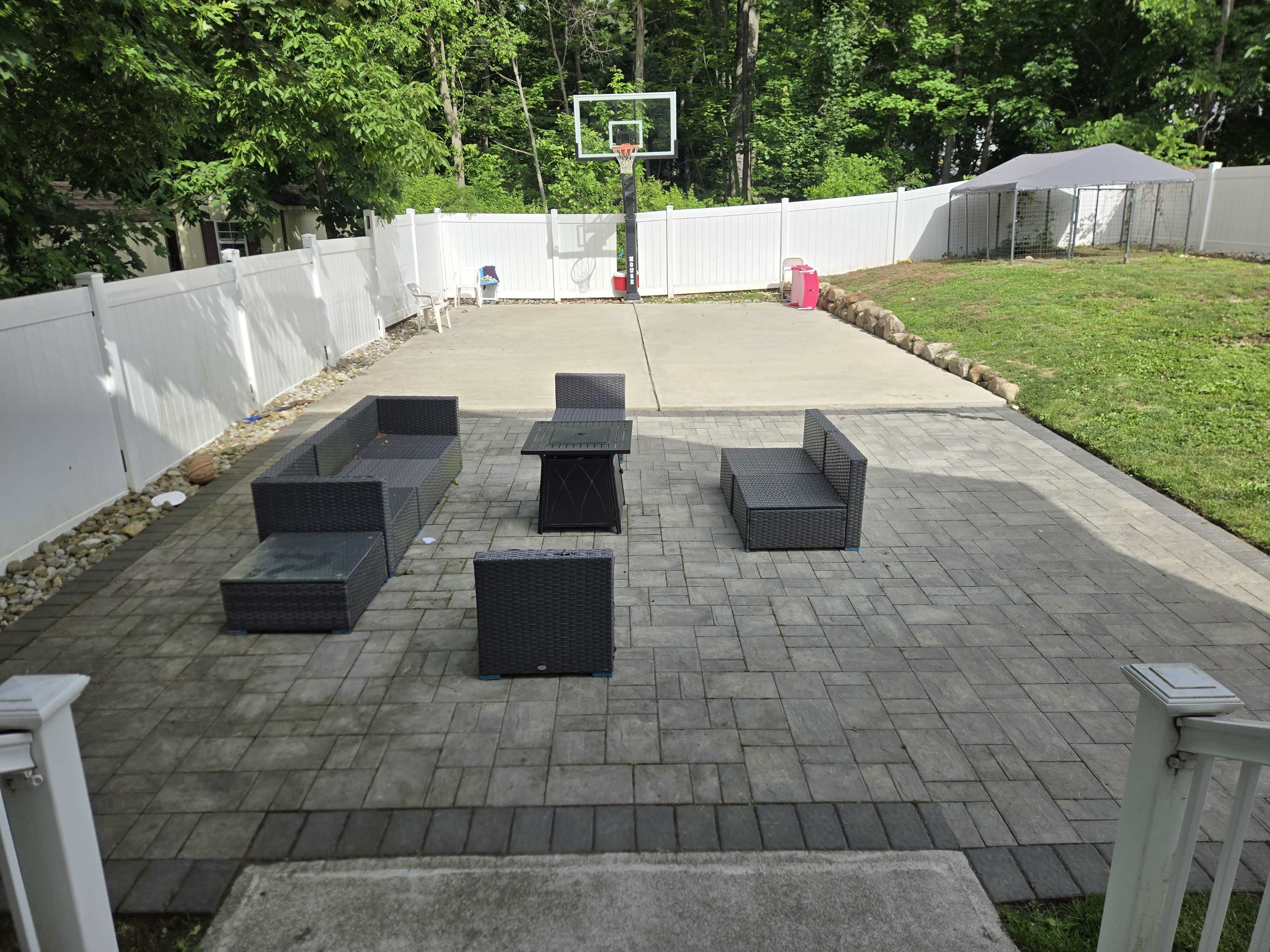 A backyard patio features a seating area with several outdoor couches and a coffee table, alongside a basketball hoop and a grassy area.