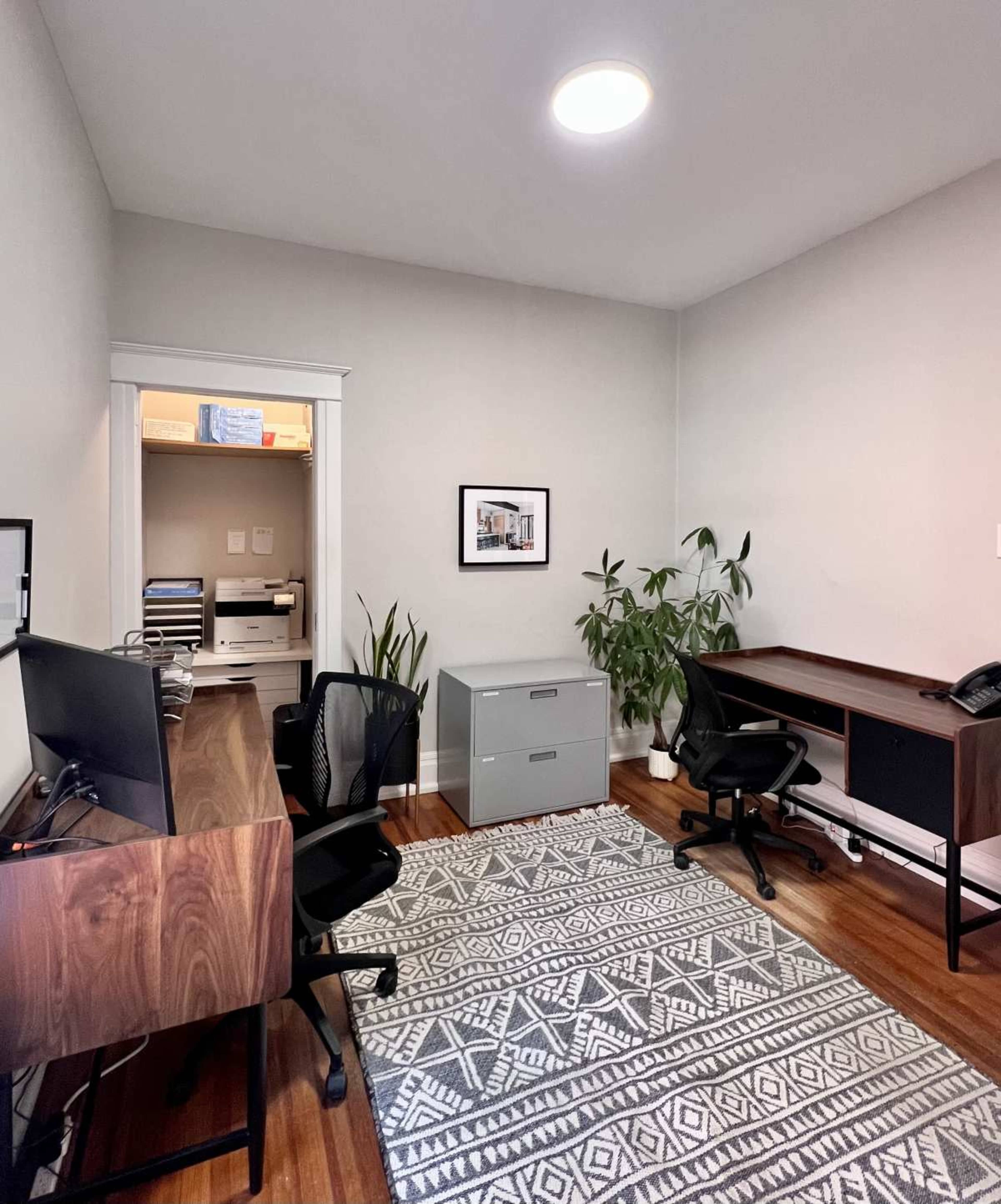 The image shows a tidy office space with two desks, a filing cabinet, a plant, and a door leading to a closet.