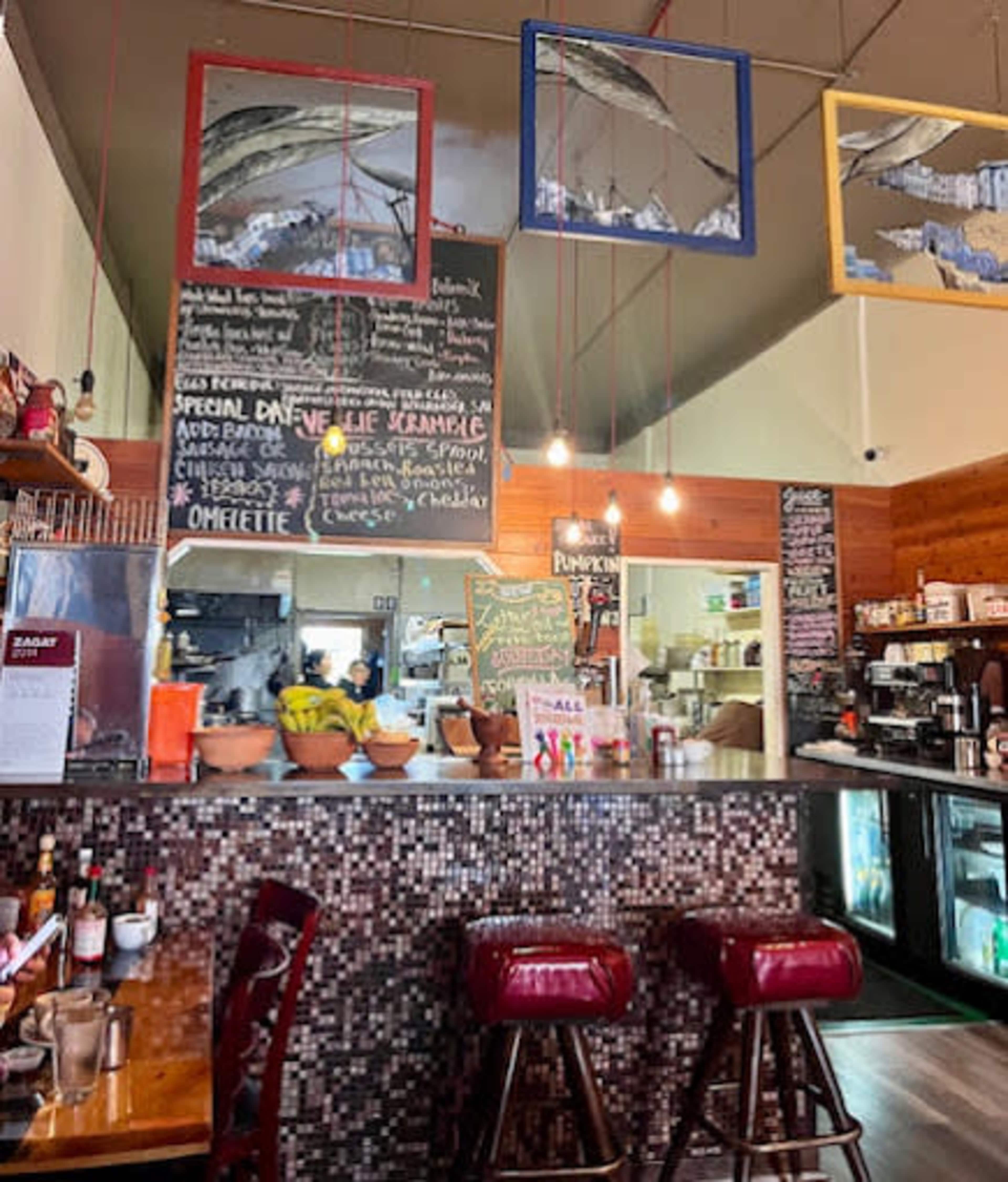 The image shows a cozy café interior with a counter displaying menu items on a chalkboard and framed artwork hanging above.