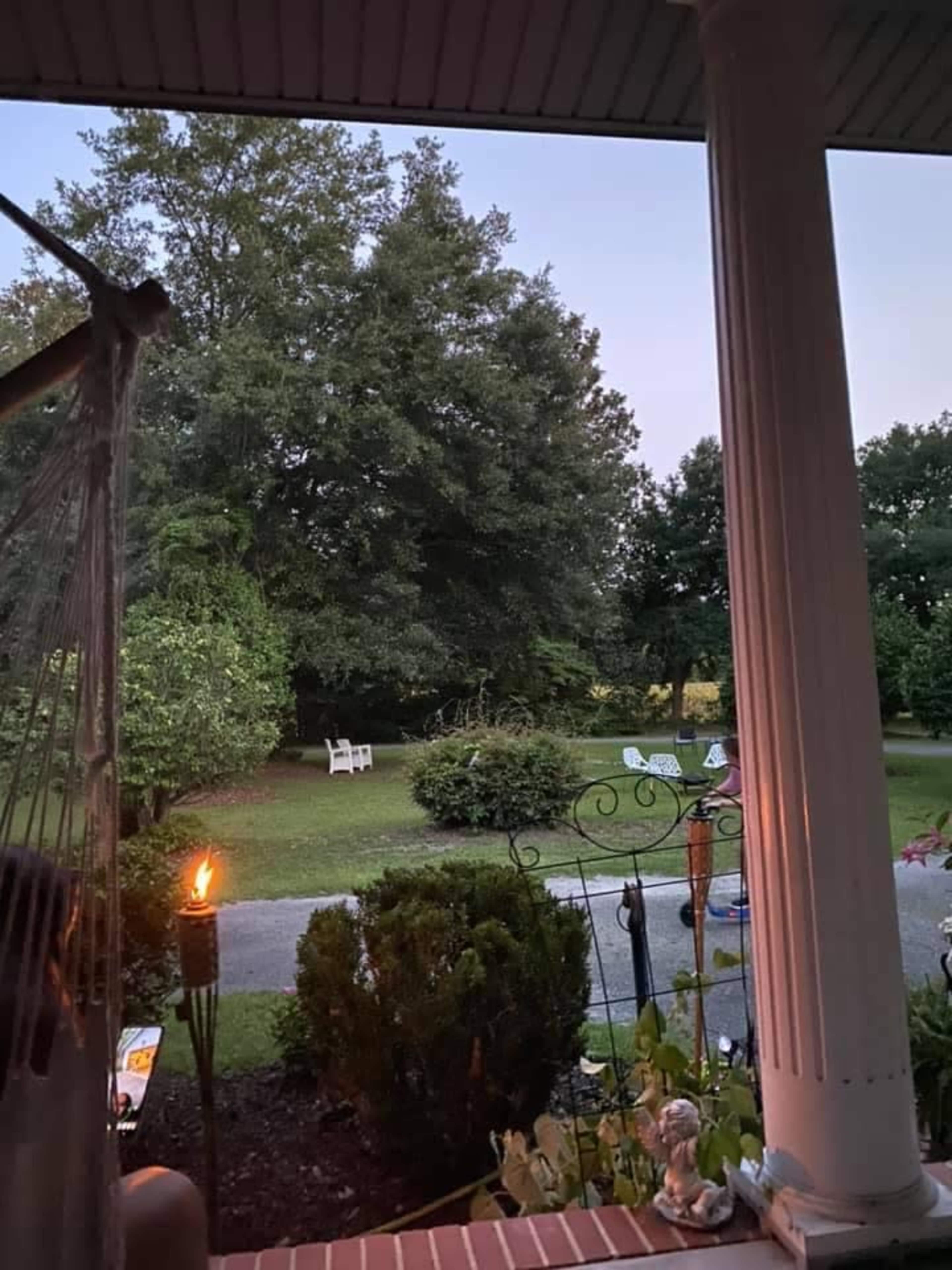 The view from a porch shows a garden with trees, a few chairs, and a lit torch in the foreground.