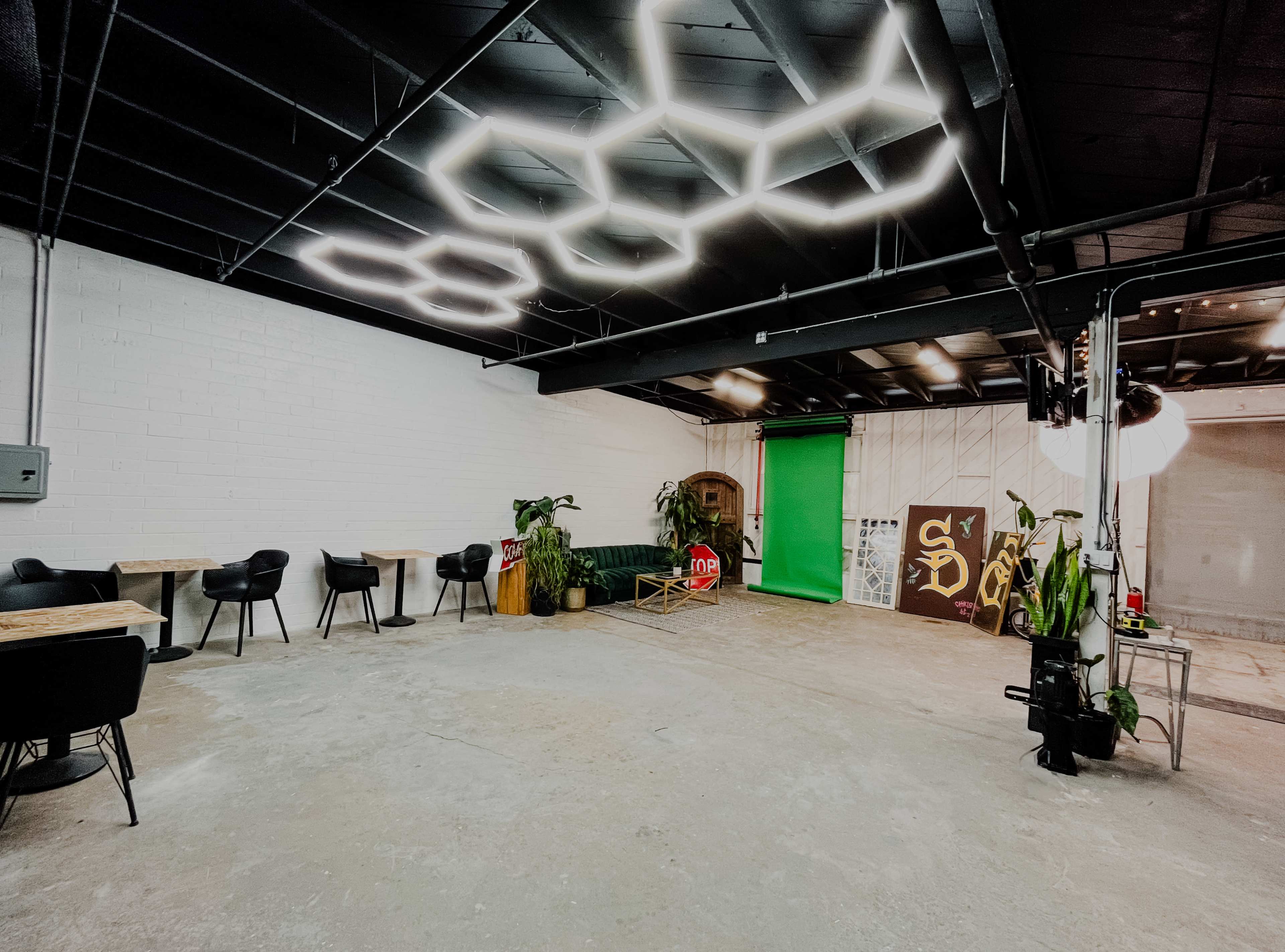 The image shows a spacious, well-lit interior with modern seating, greenery, and a backdrop for photography or videography on one side.