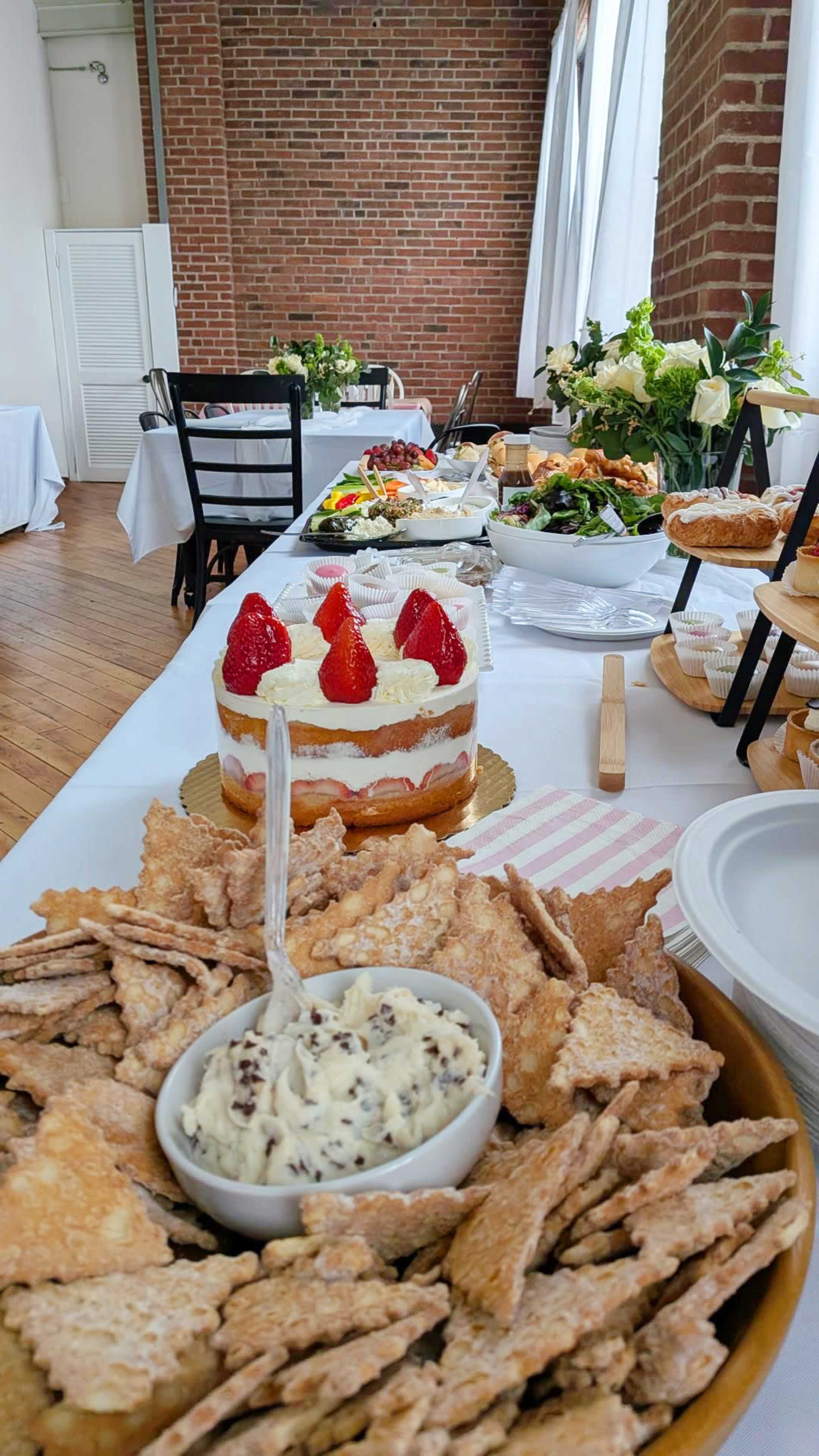 A long table is set with various dishes, including a cake topped with strawberries and a bowl of dip surrounded by crackers.