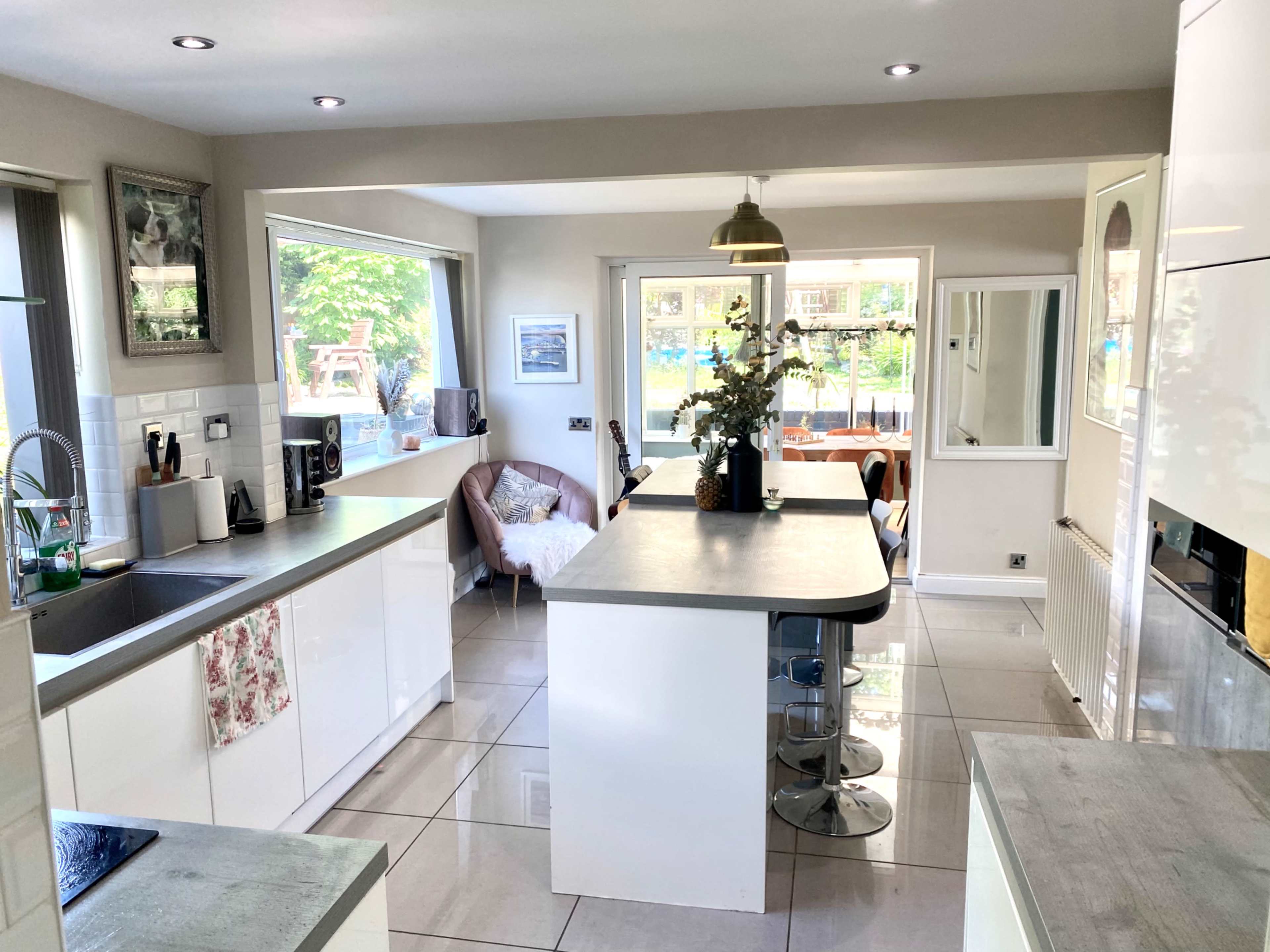 A modern kitchen features sleek white cabinetry, a large island with a gray countertop, and a view of a sunny outdoor seating area through the windows.