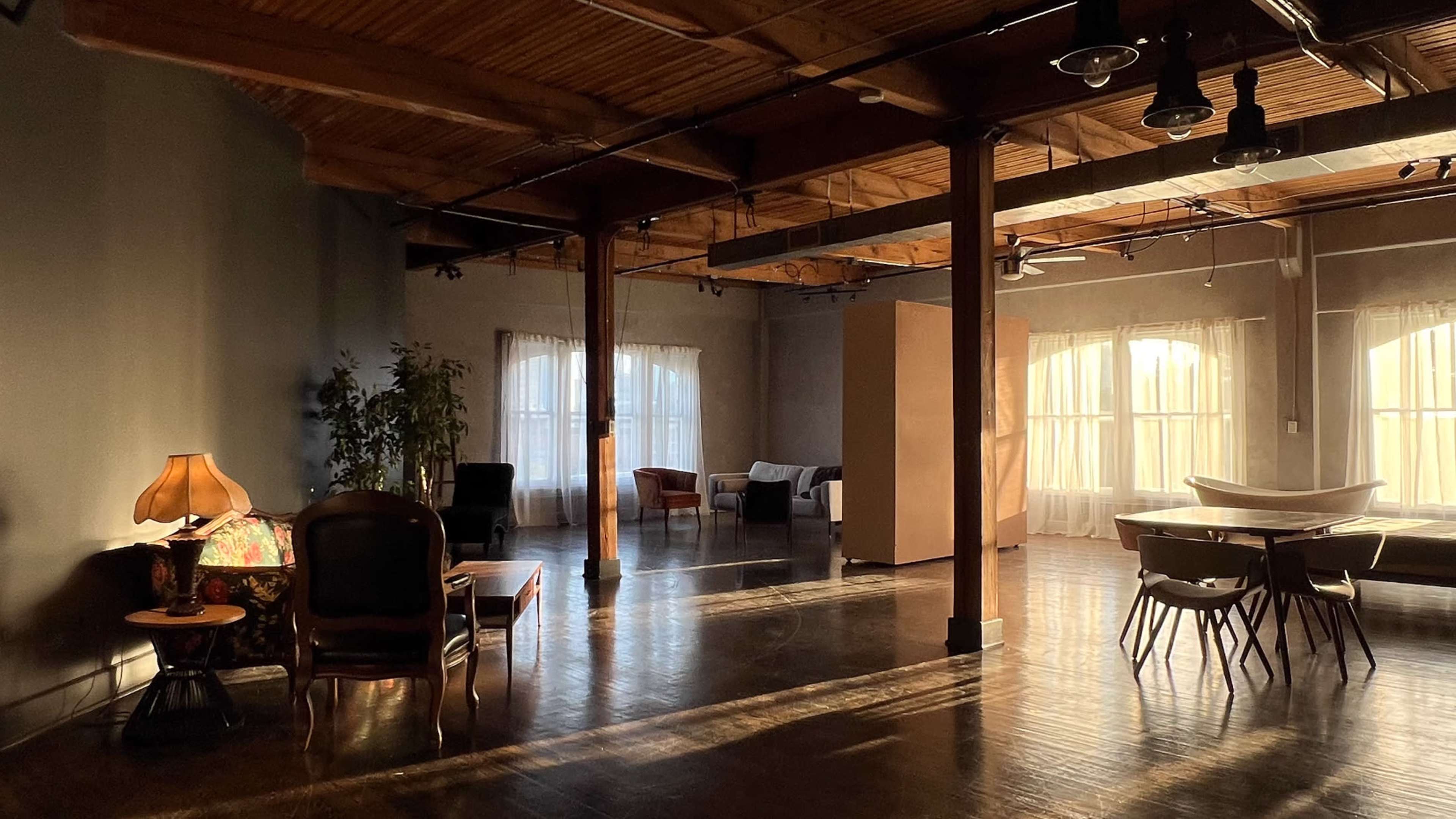 The image shows a spacious, well-lit interior with wooden beams, various seating areas, and large windows allowing sunlight to enter.